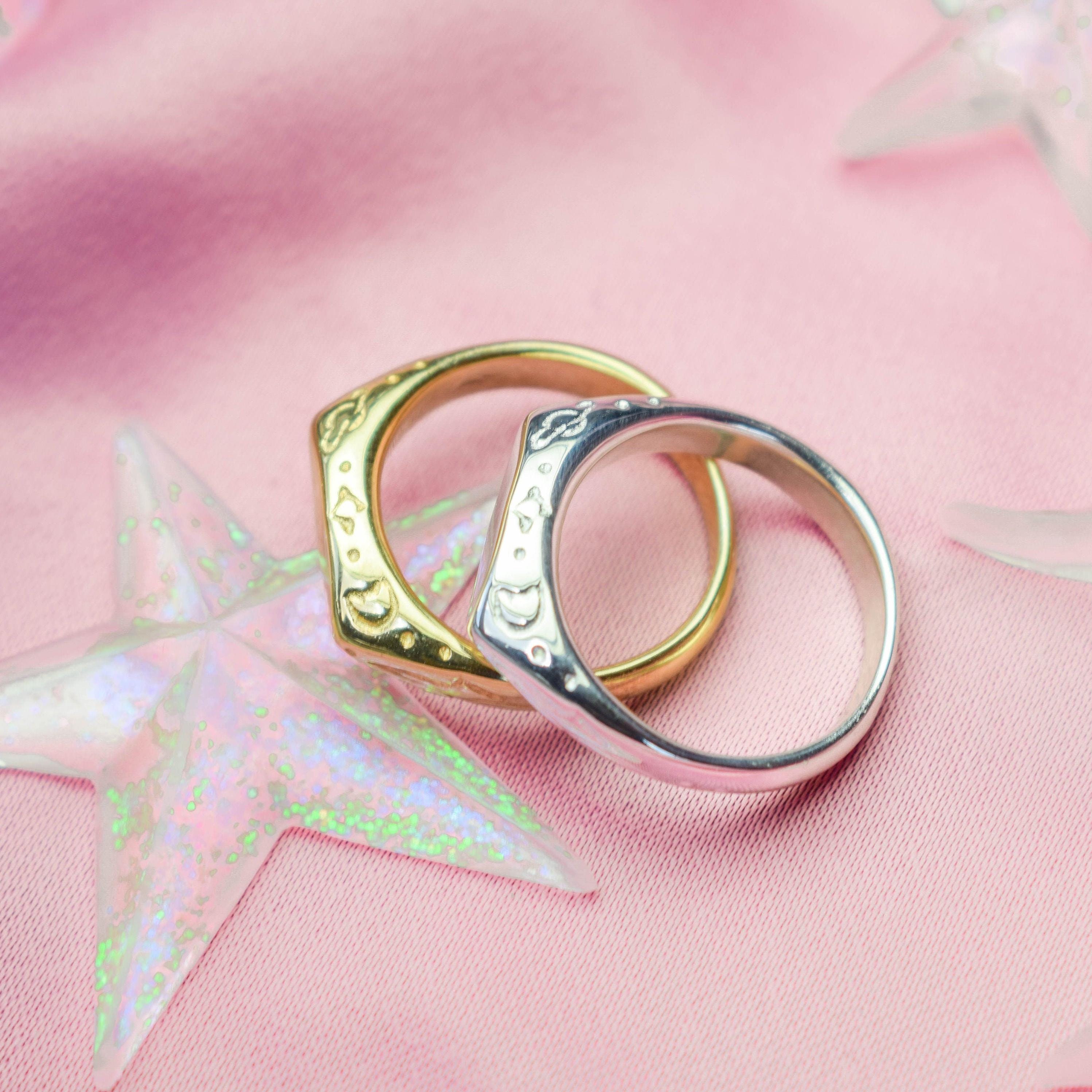 Two dainty signet rings, one with a gold tone and the other in silver, placed on a pink background with star patterns.