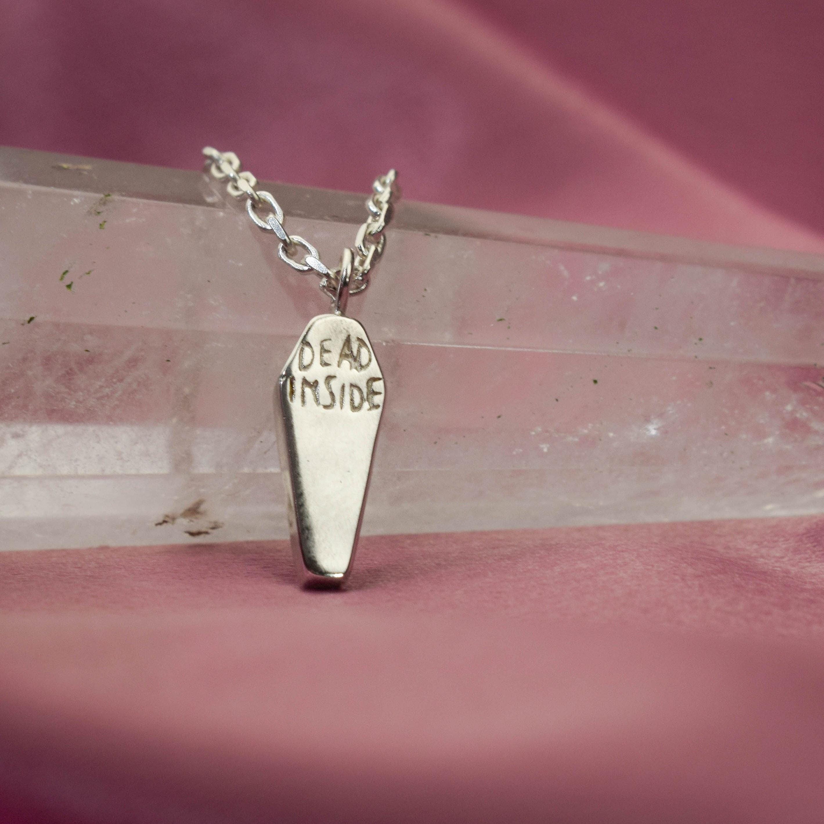 A silver-colored minimalist coffin-shaped necklace with the inscription 'DEAD INSIDE' on a pink background.