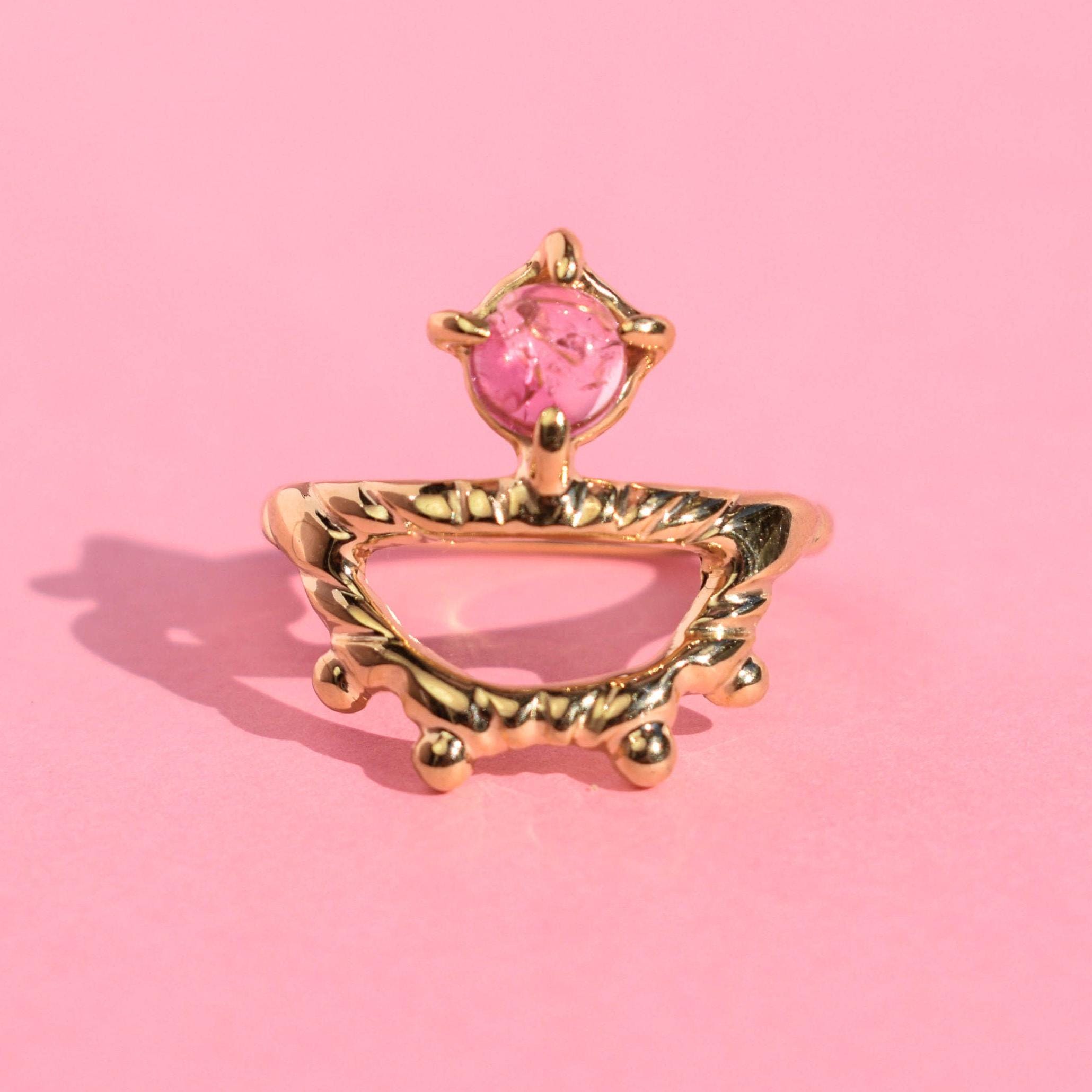 A gold-colored ring with an intricate design featuring a pink tourmaline gemstone on a pink background.