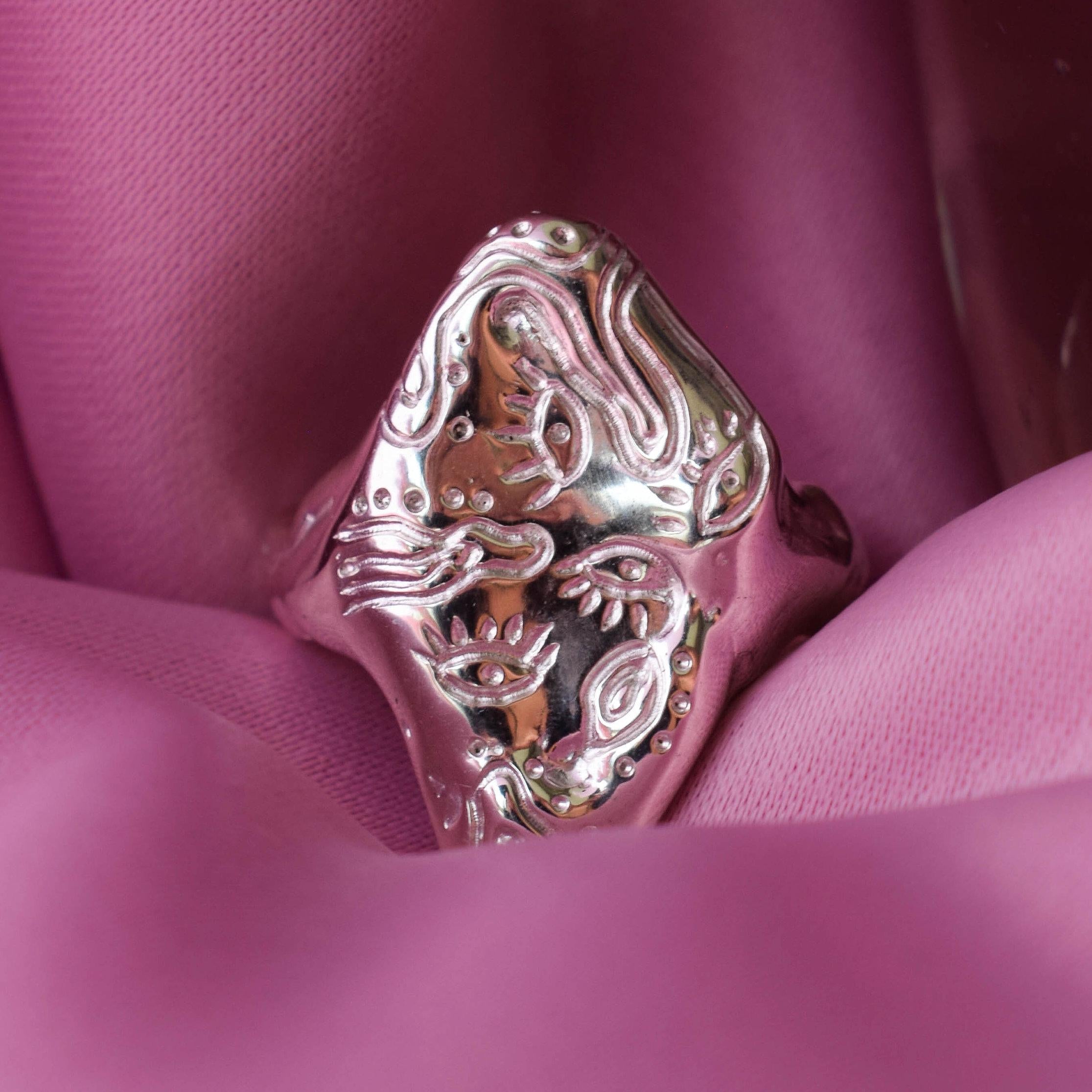 A sterling silver ring with a detailed hand-carved design resembling eyes, displayed on a pink fabric background.