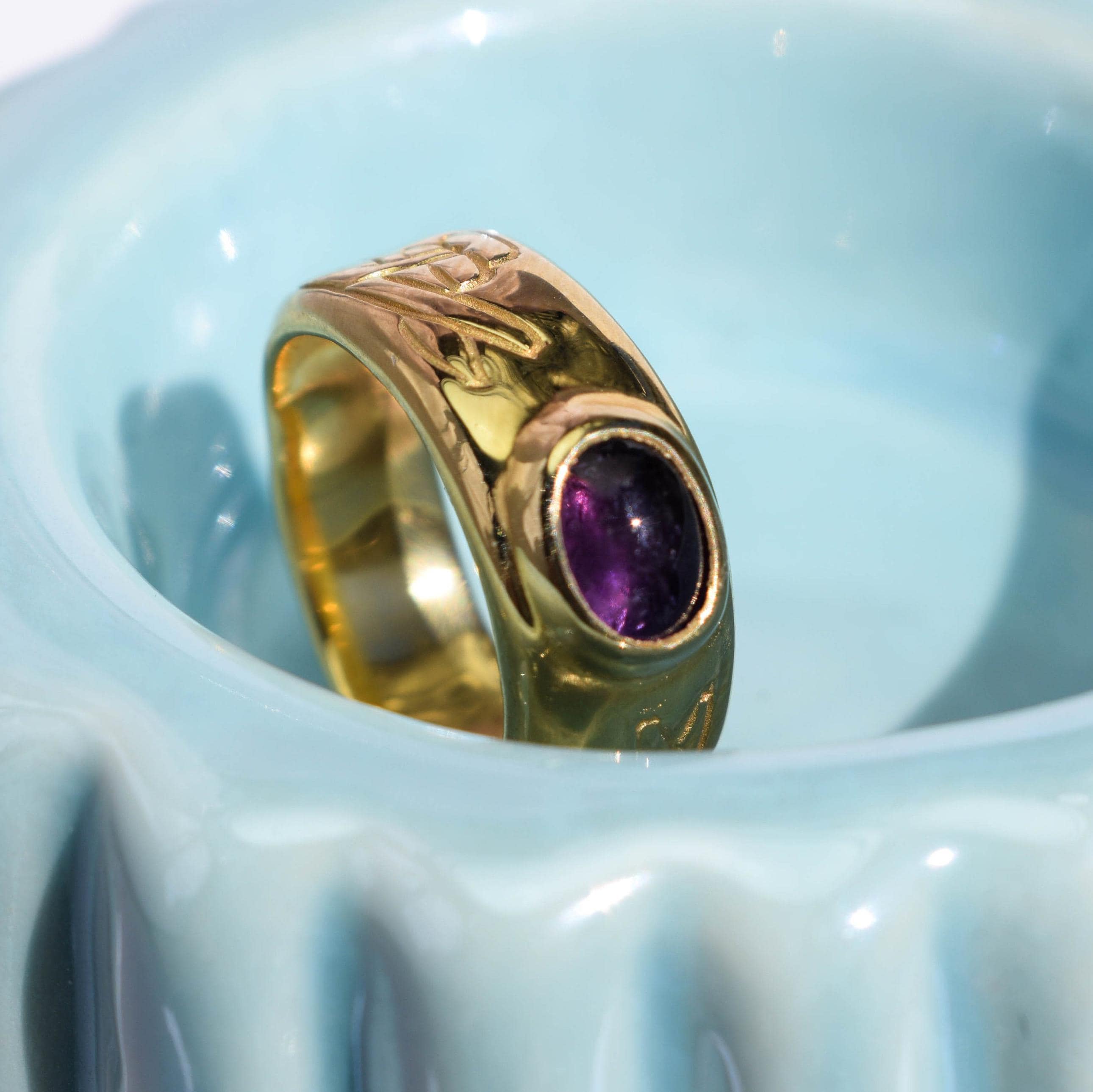 A gold-toned ring with a purple gemstone, featuring an old school tattoo-inspired design, displayed in a blue ceramic dish.