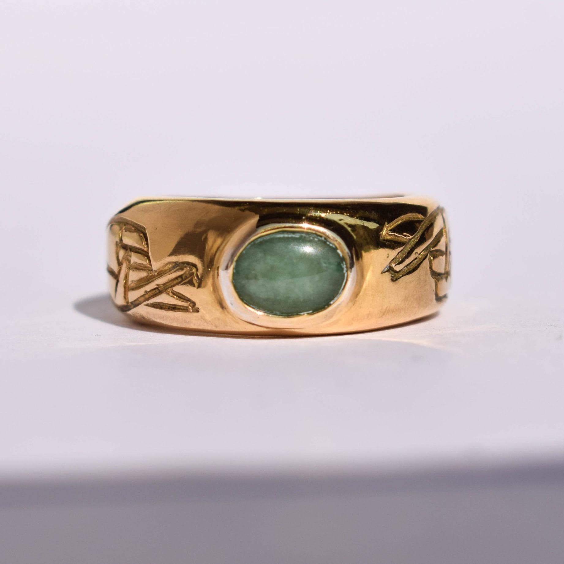 A gold-colored ring with a green gemstone in the center, featuring old school tattoo-inspired design patterns on the band.
