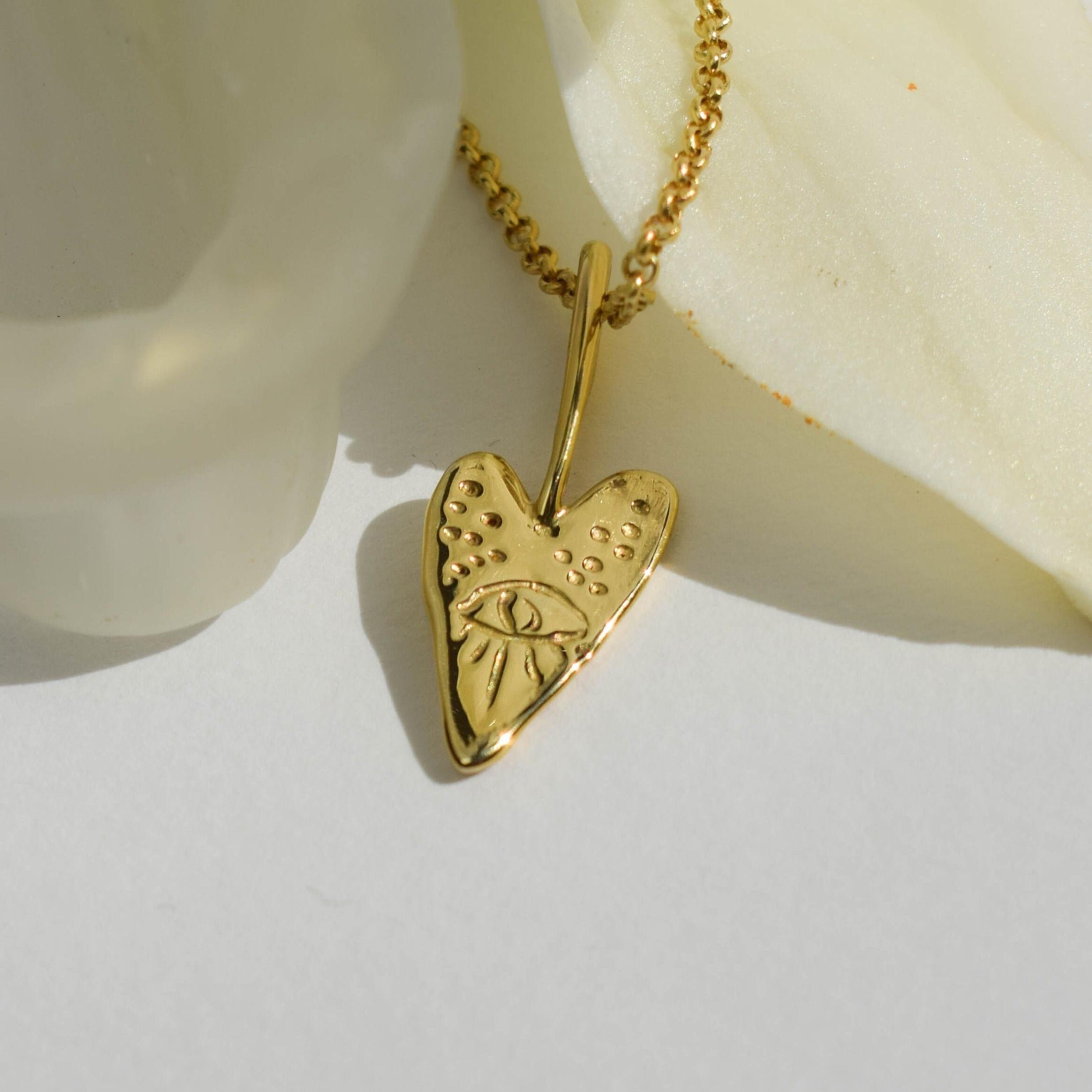 A gold-tone necklace featuring a pendant with a heart shape and an eye in the center, displayed on a white surface with a flower.