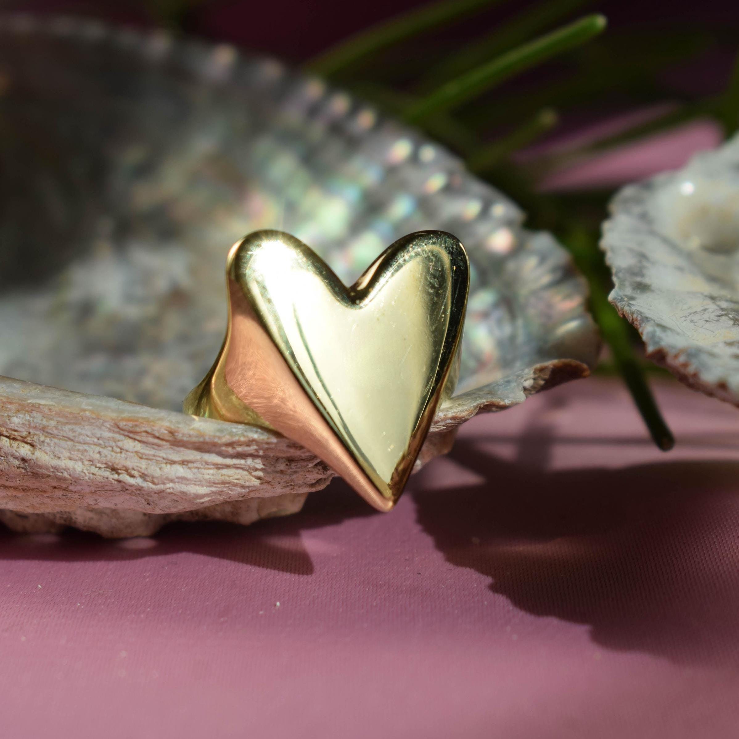 A gold-colored heart-shaped signet ring placed on a seashell, with a pink background and green foliage in the background.