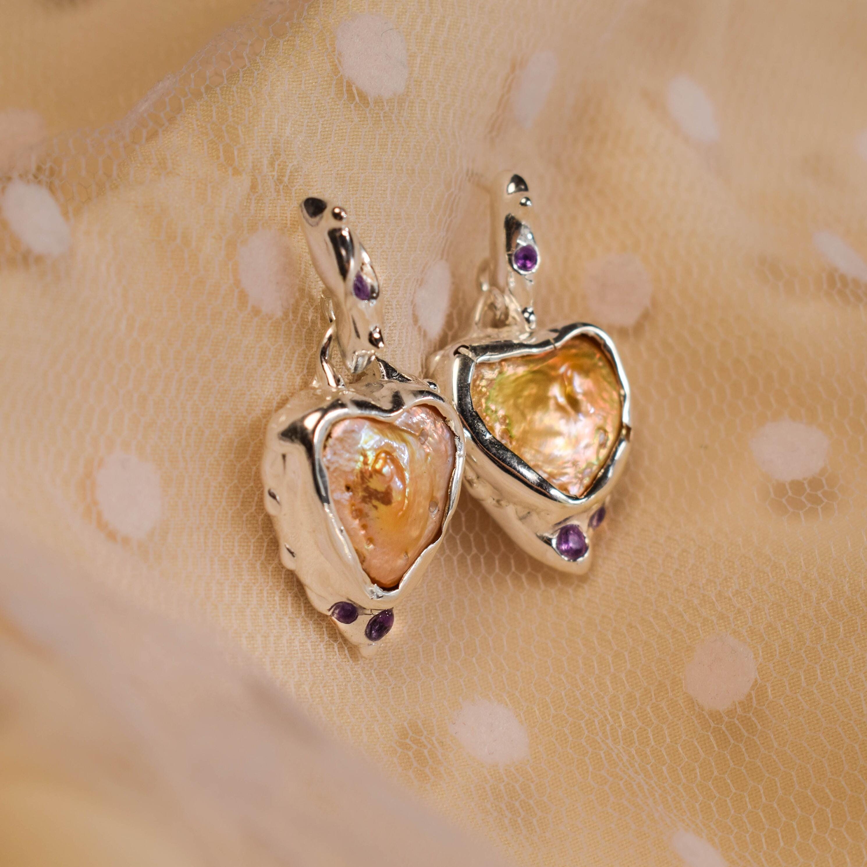 A pair of heart-shaped statement earrings with mother of pearl hearts and amethyst accents, displayed against a beige textured background.