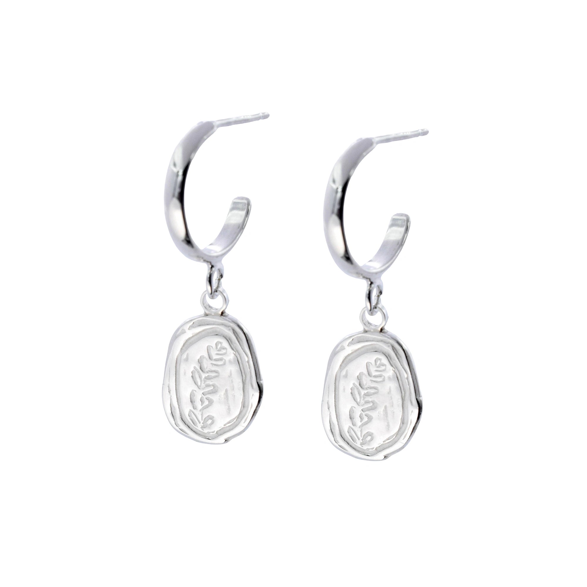 A pair of silver huggie hoop earrings with dangling oval charms featuring an engraved botanical leaf pattern, shown against a plain white background.