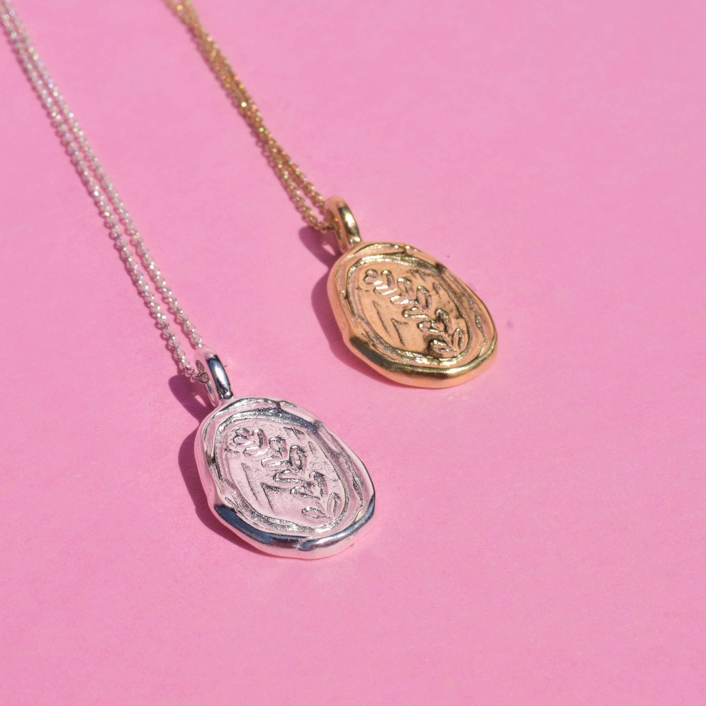 Two botanical pendant necklaces, one with a silver tone and the other with a gold tone, both featuring a carved leaf design on a pink background.