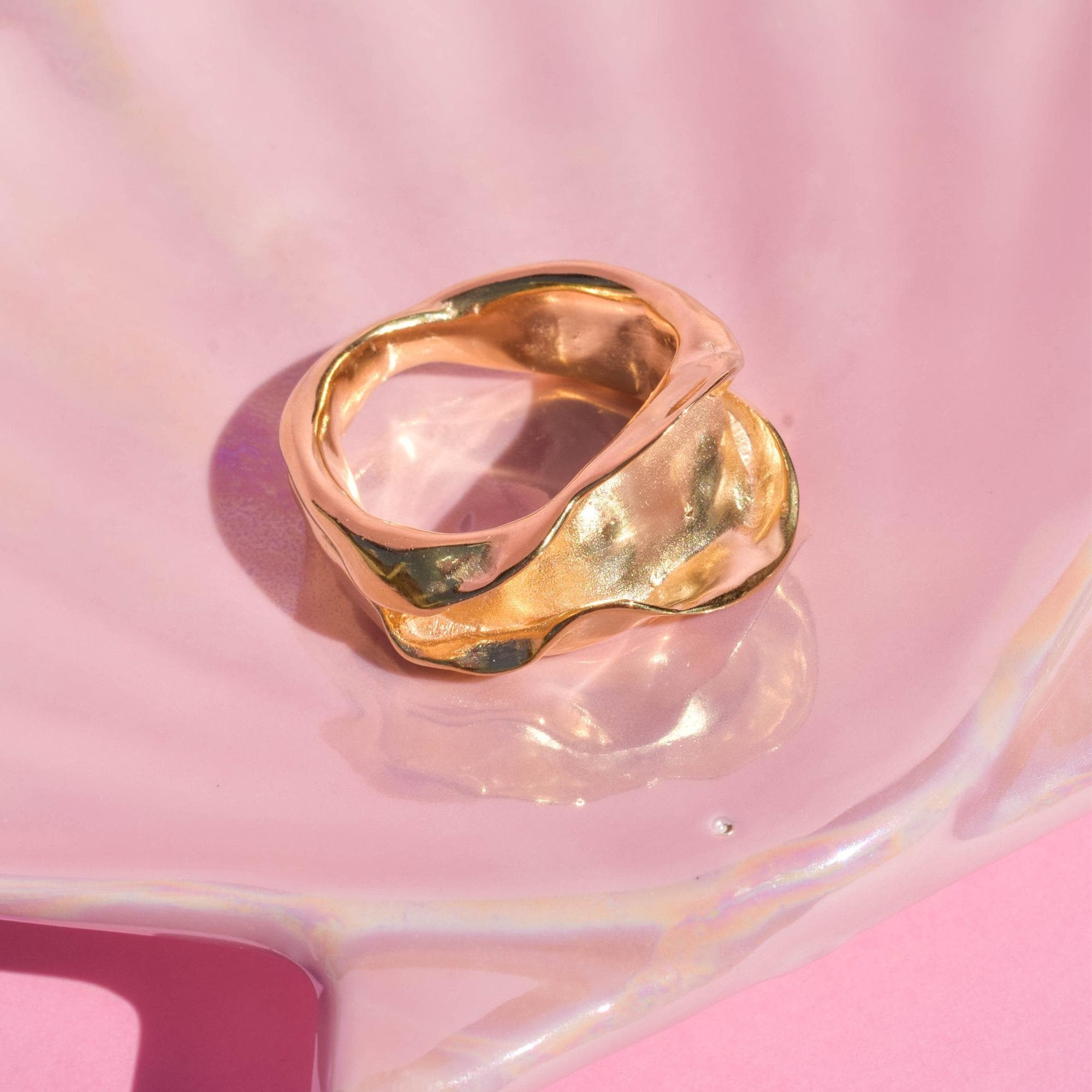 A gold-colored minimalistic ring with a folded design, displayed on a pink surface.