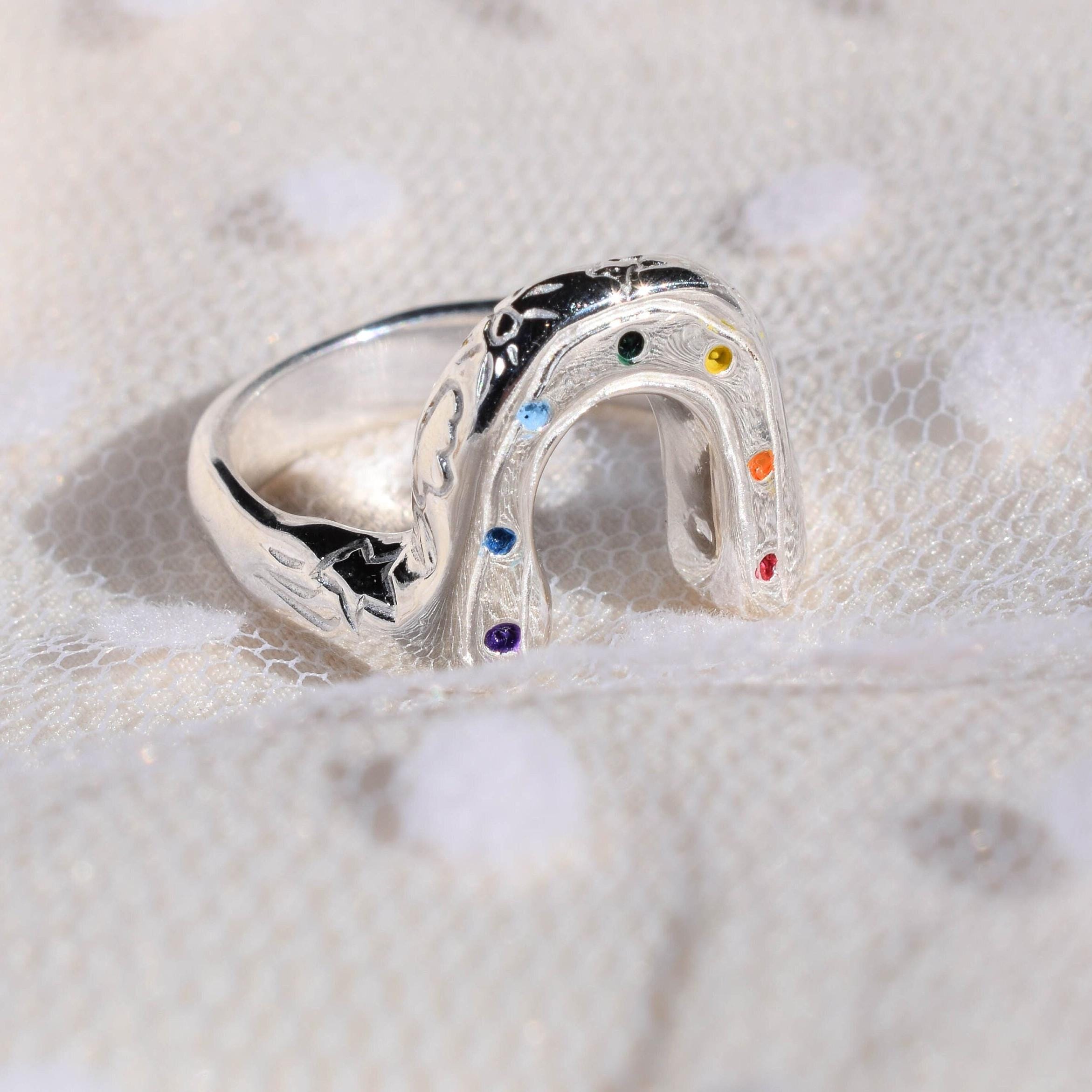 A silver-colored ring with a rainbow design, featuring multicolored acrylic paint and decorative elements like clouds, suns, moons, and stars on the sides, displayed on a white textured background.