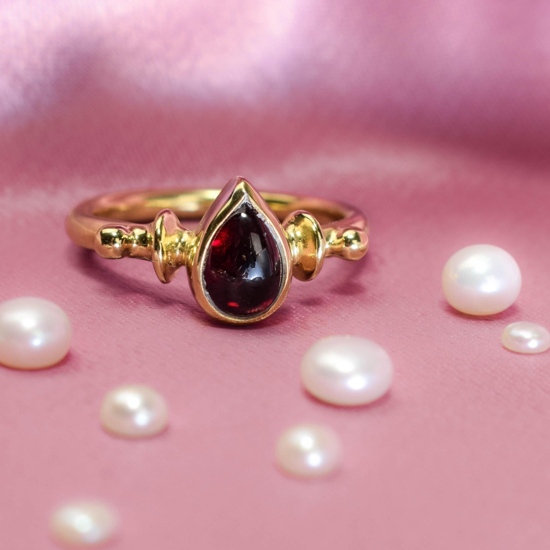 A teardrop-shaped red garnet solitaire ring with gold-tone detailing, displayed against a pink background with scattered pearls.