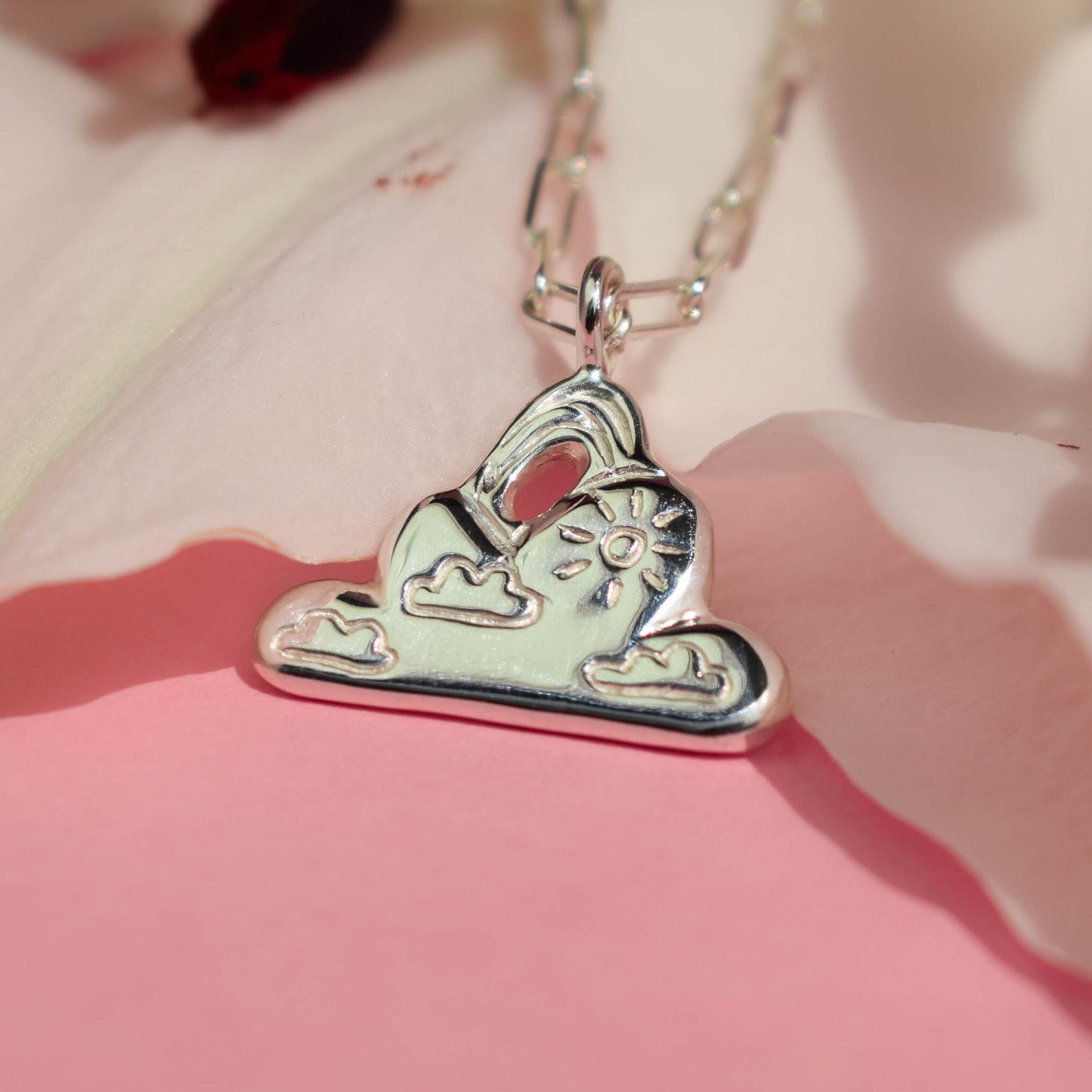 A silver cloud-shaped pendant necklace with a sun and rainbow details, displayed against a pink background with a flower.