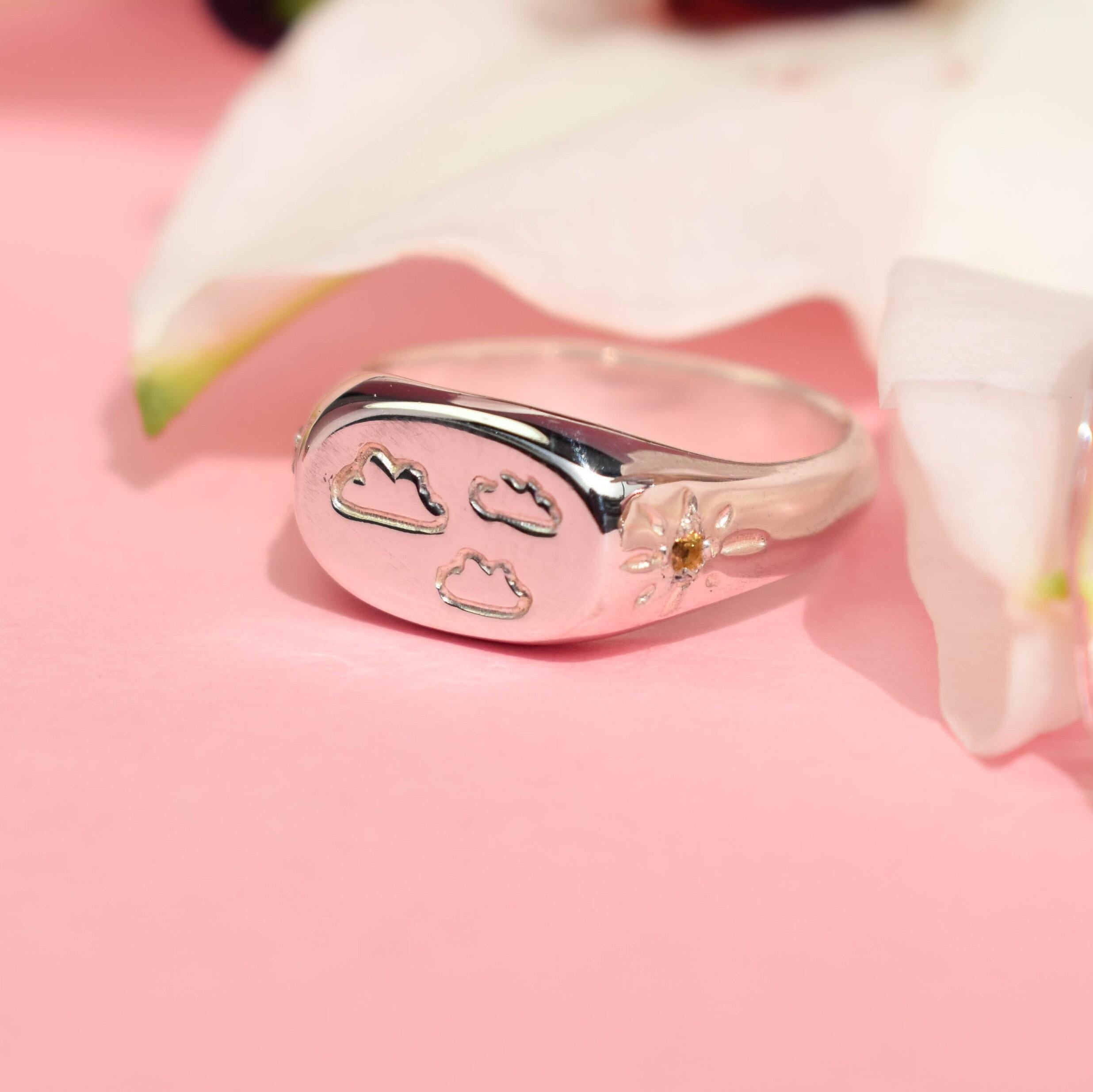A silver signet ring with a design of clouds and a yellow sapphire sun, displayed on a pink surface with a white flower in the background.