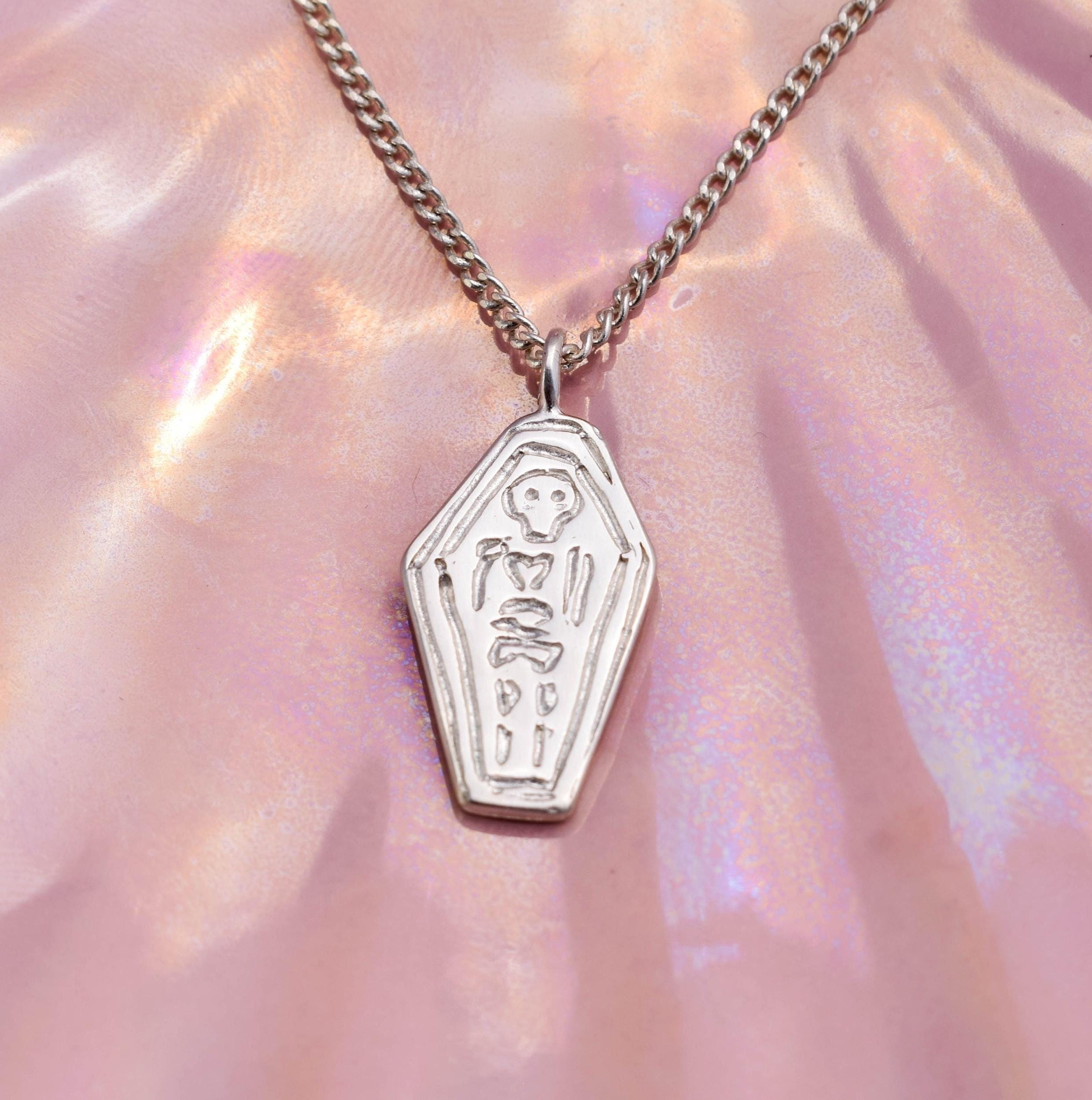 A silver-colored necklace featuring a pendant in the shape of a coffin with a skeleton design, displayed against a pink, translucent background.