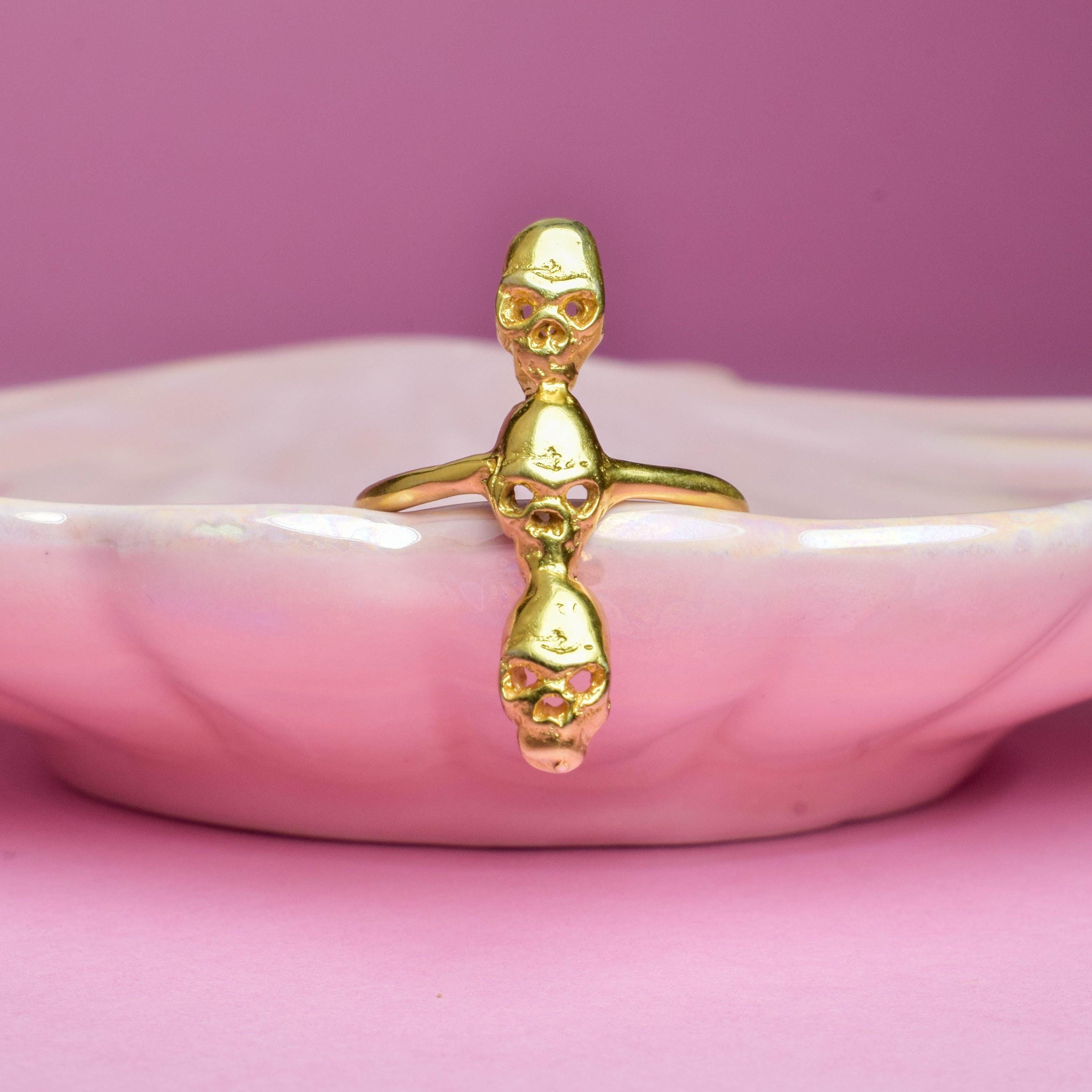 A gold-colored ring with three skull designs forming a totem-like structure, displayed on a white dish against a pink background.