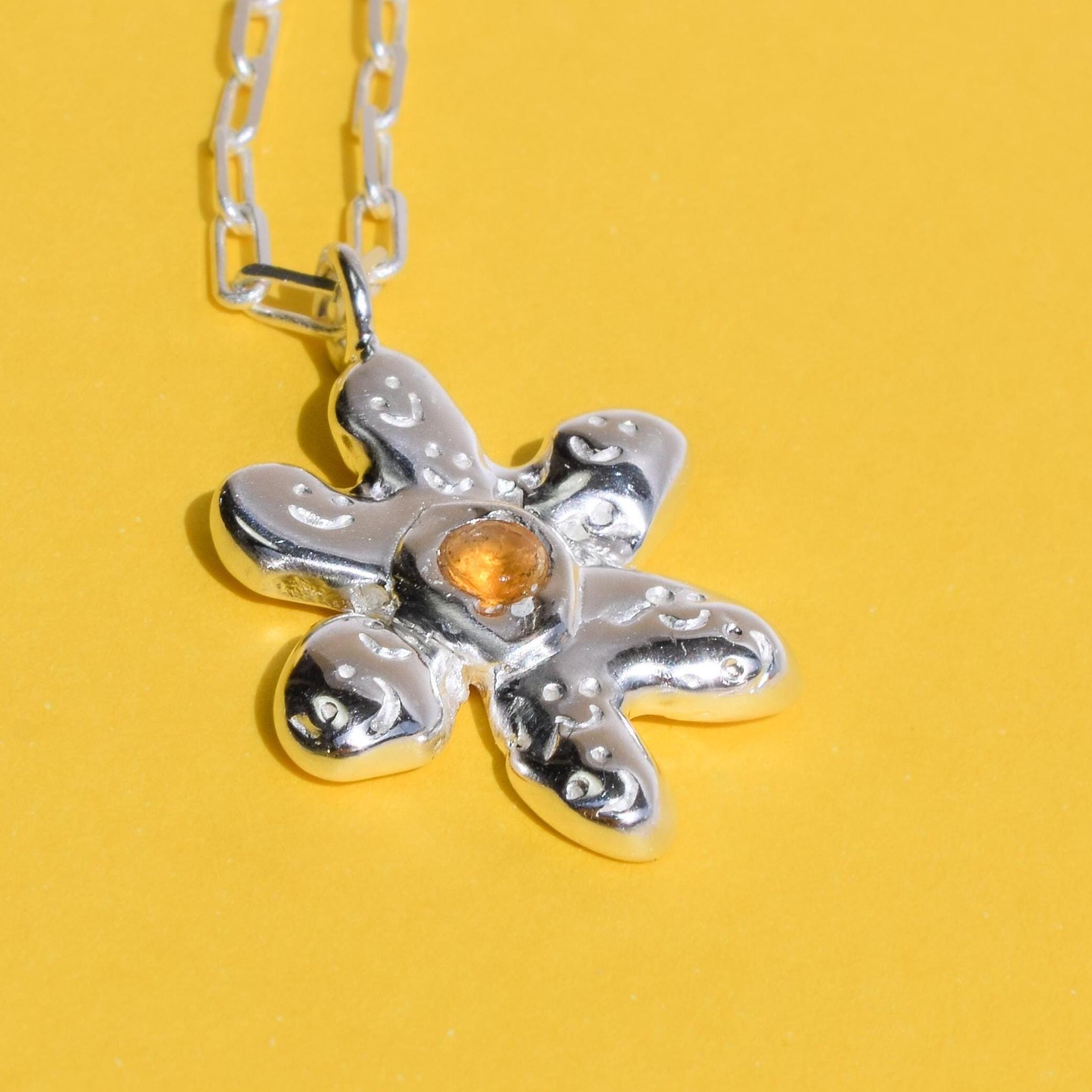 A silver daisy-shaped necklace with a yellow citrine gemstone in the center, displayed against a yellow background.