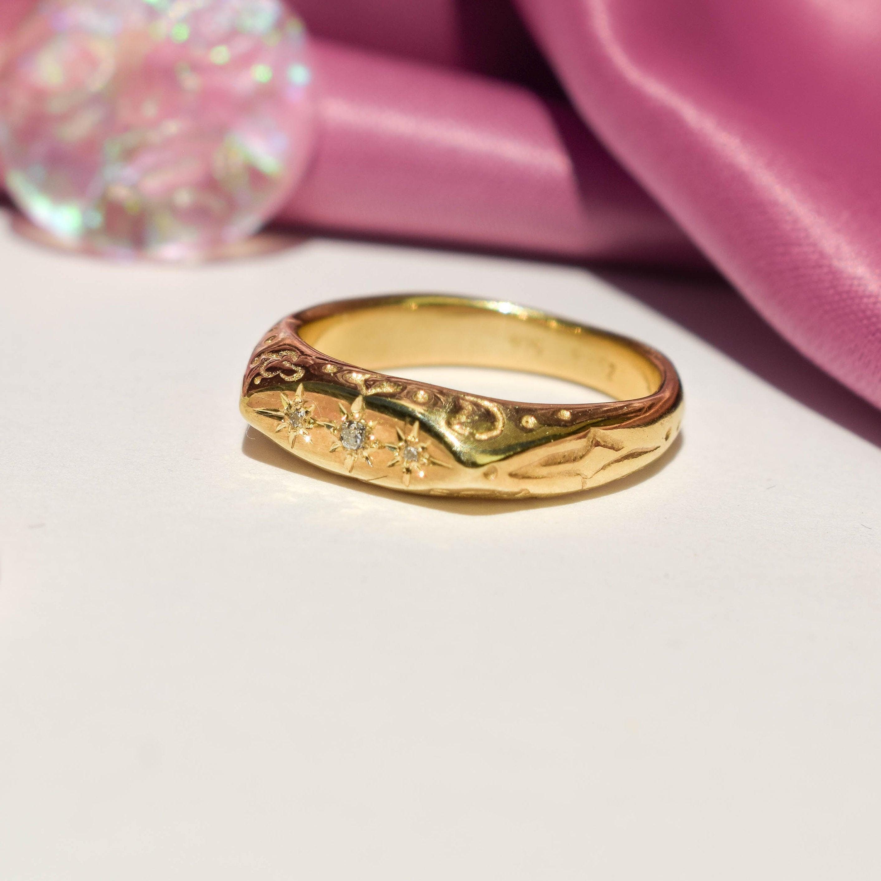 A gold-colored ring with clear cubic zirconia crystals arranged in a starburst pattern, displayed on a white surface with a pink item in the background.