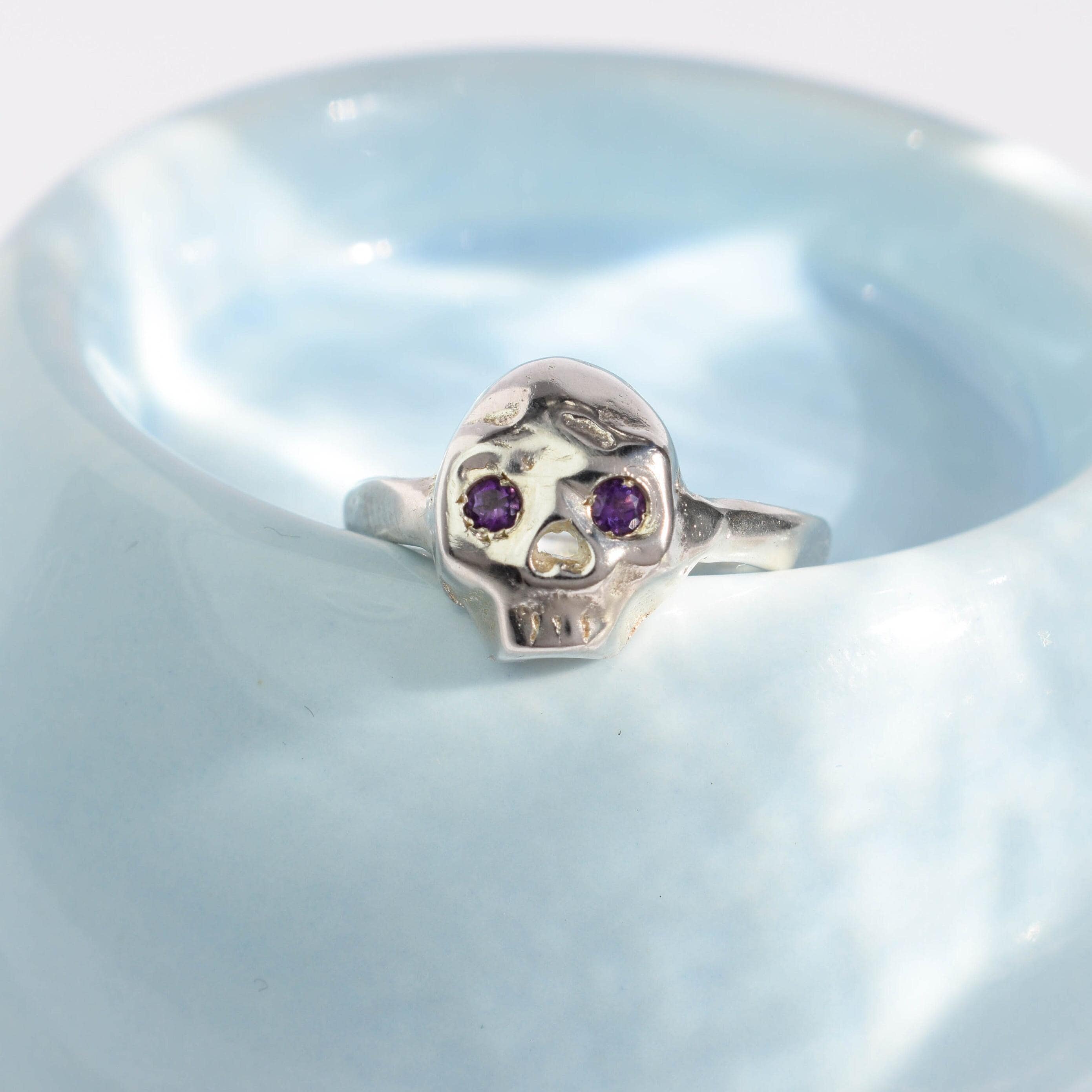 A sterling silver skull ring with two purple amethysts set in the eye sockets, displayed on a blue textured surface.