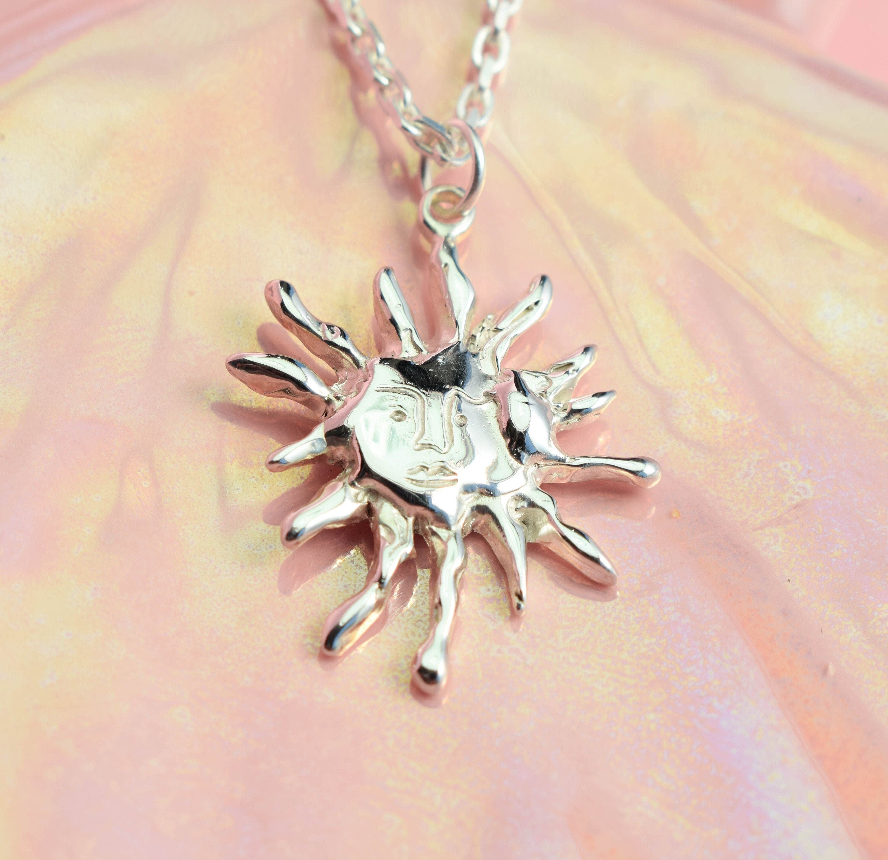 A shiny, silver-tone sun necklace pendant on a chain, displayed against a pink background.