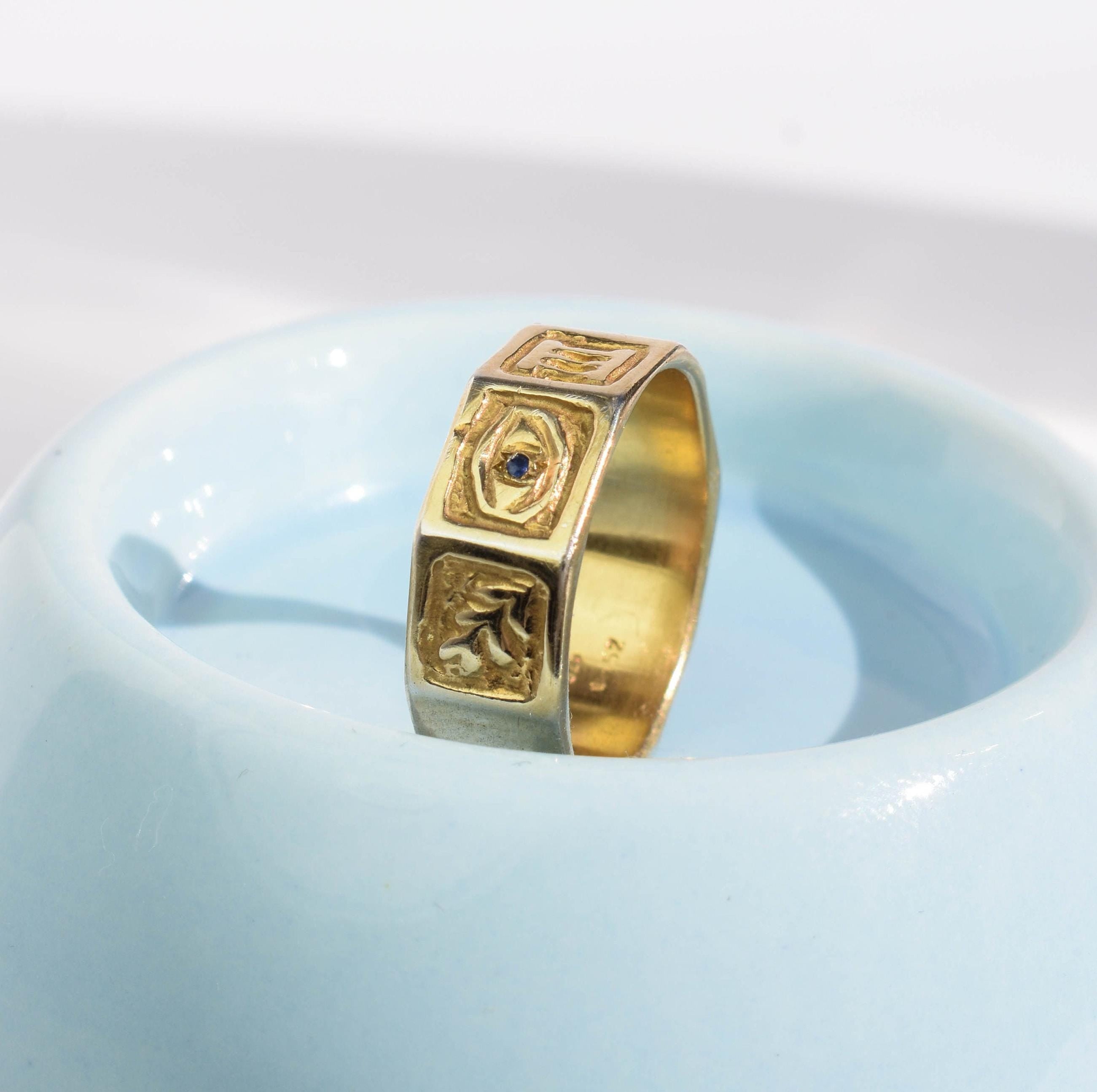 A gold-colored band ring with intricate designs, including an evil eye and a branch with leaves, placed on a blue dish.
