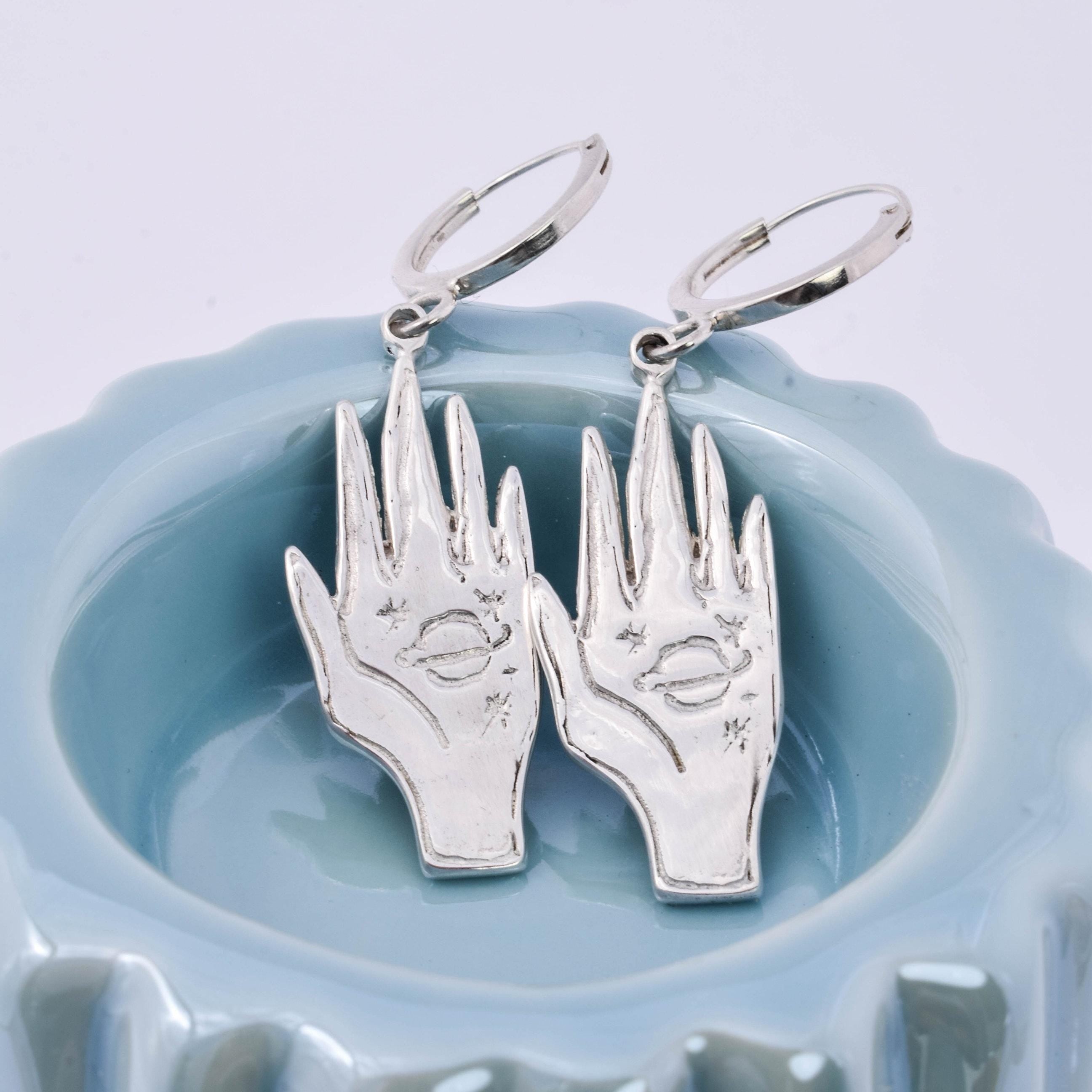 A pair of silver charm hoop earrings designed to resemble hands holding a small universe, displayed on a blue dish.