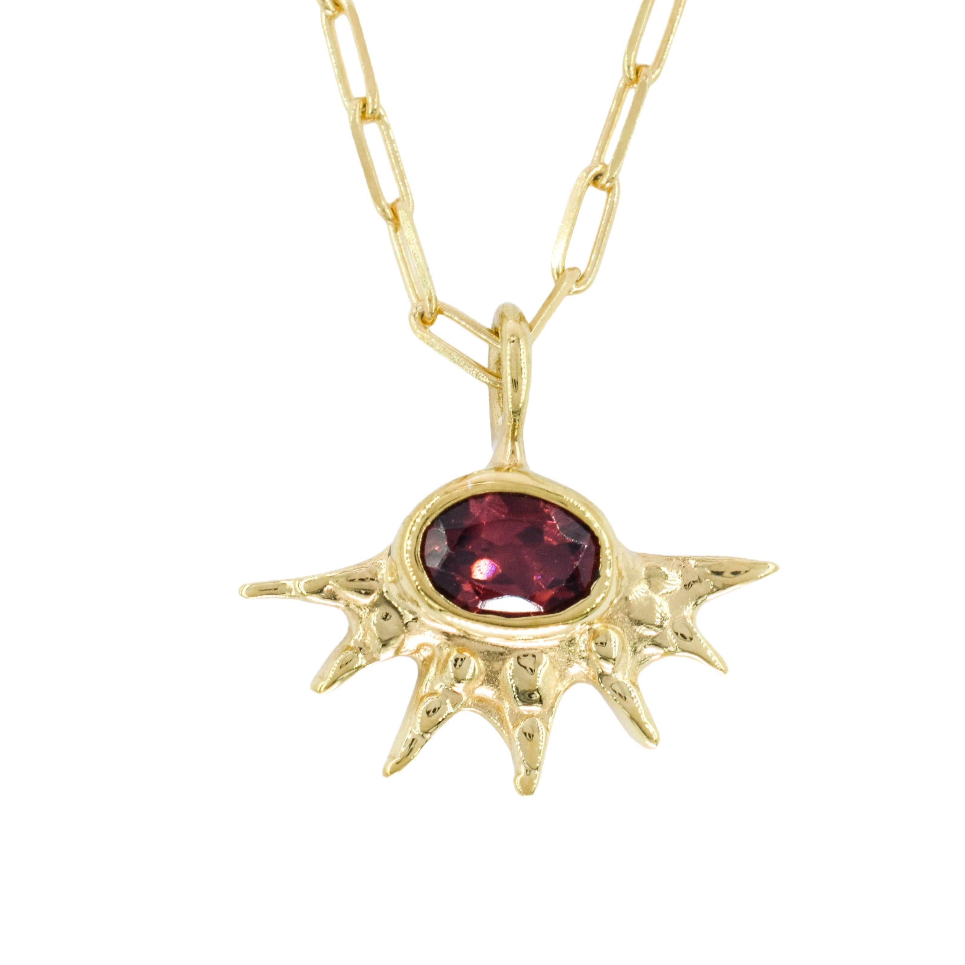 A gold paperclip chain necklace with a pendant shaped like a stylized sun or eye, featuring a faceted dark red gemstone in the center, shown against a plain white background.