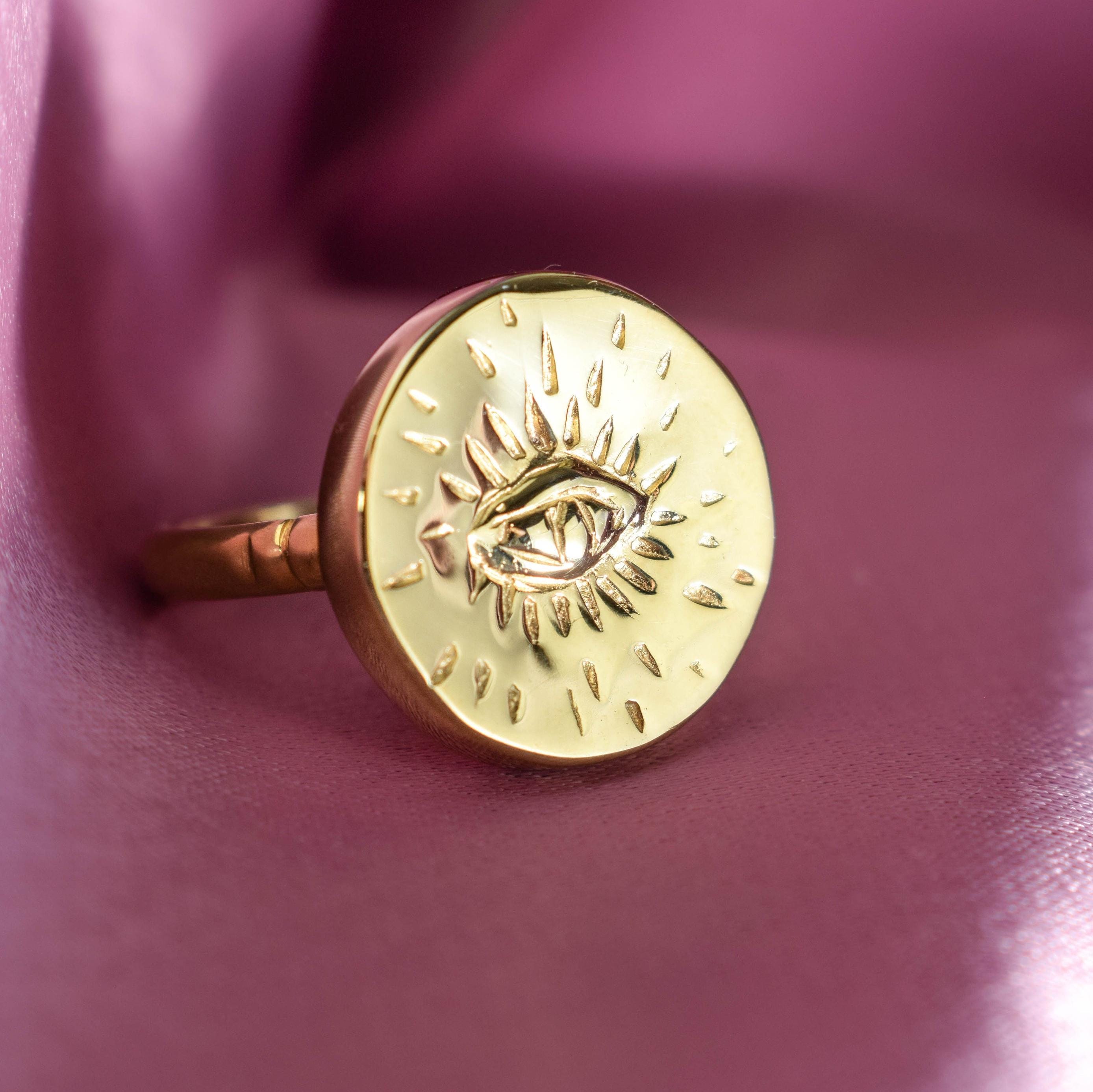 A gold-colored, round ring with intricate designs and a central eye motif, displayed against a purple background.