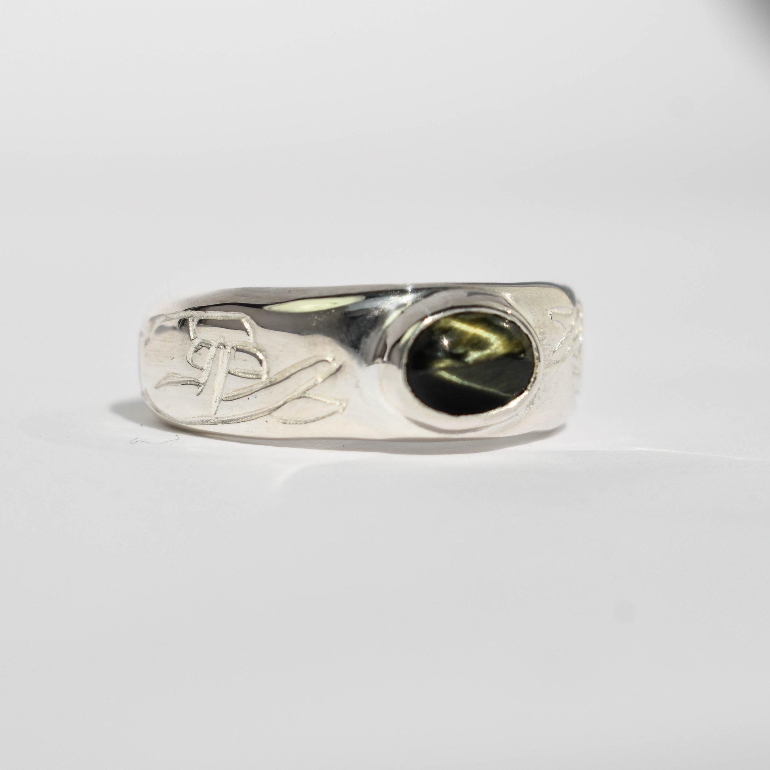 A ring with a black tiger's eye gemstone and a band featuring crossed fingers inspired by old school tattoos, set on a white background.