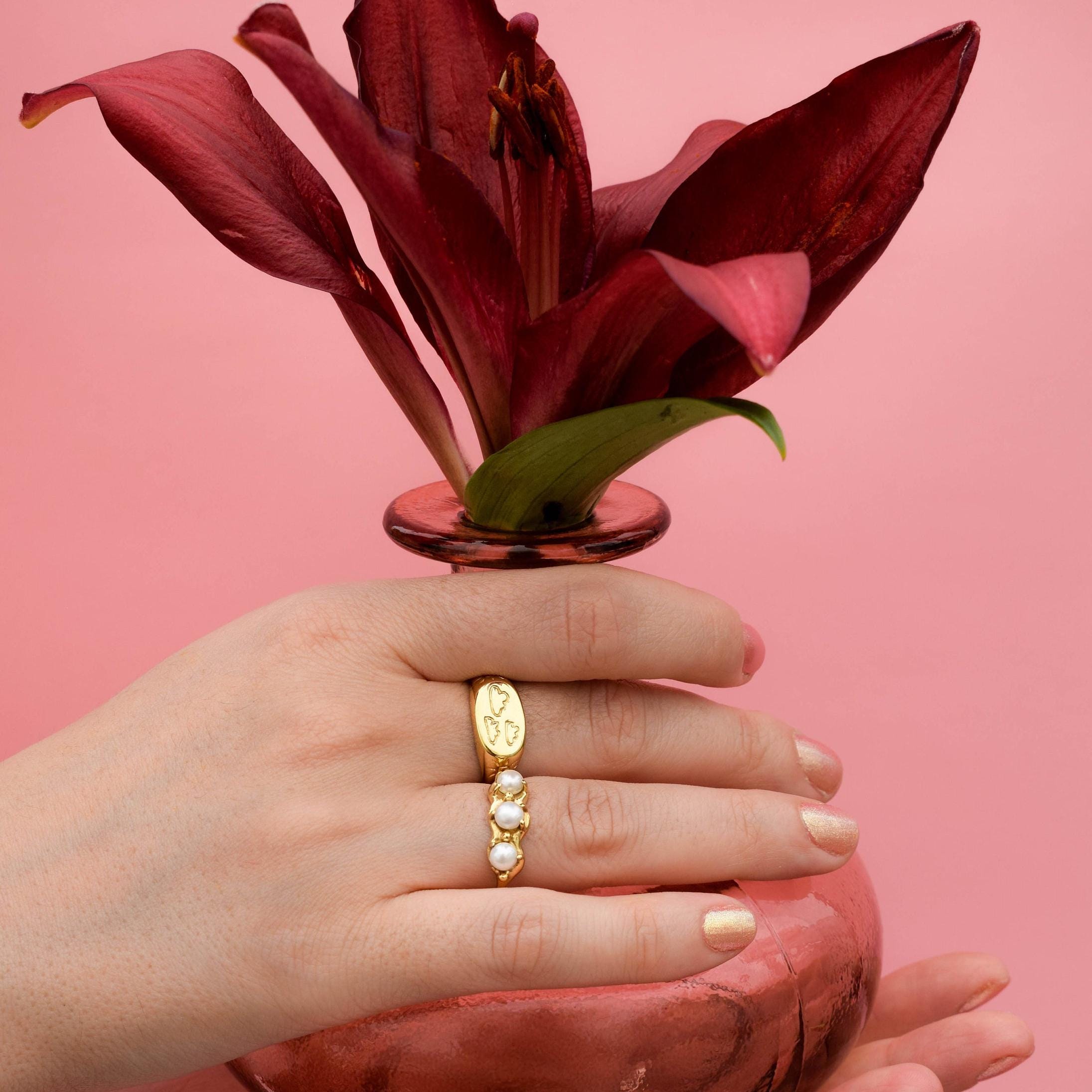 A hand wearing a gold-colored signet ring with a cloud design on a pink background, holding a red flower.