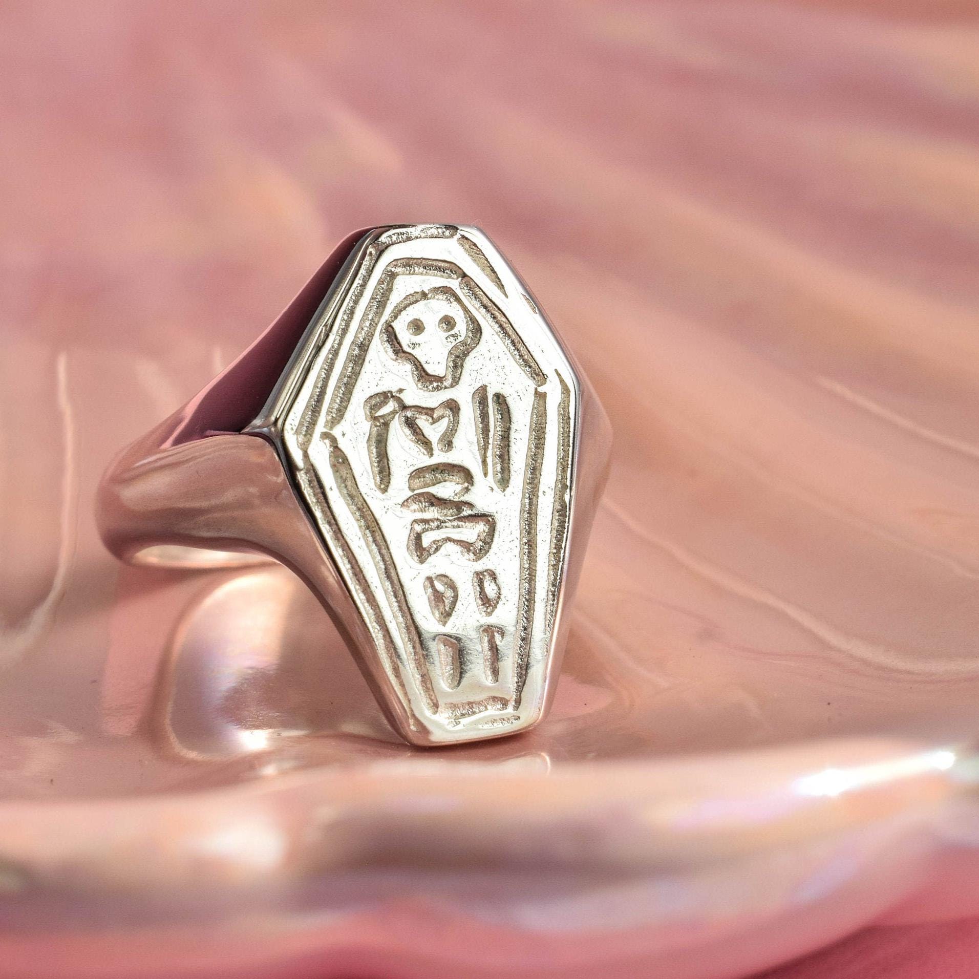 A silver signet ring with a skeleton design, placed on a pink cushion.