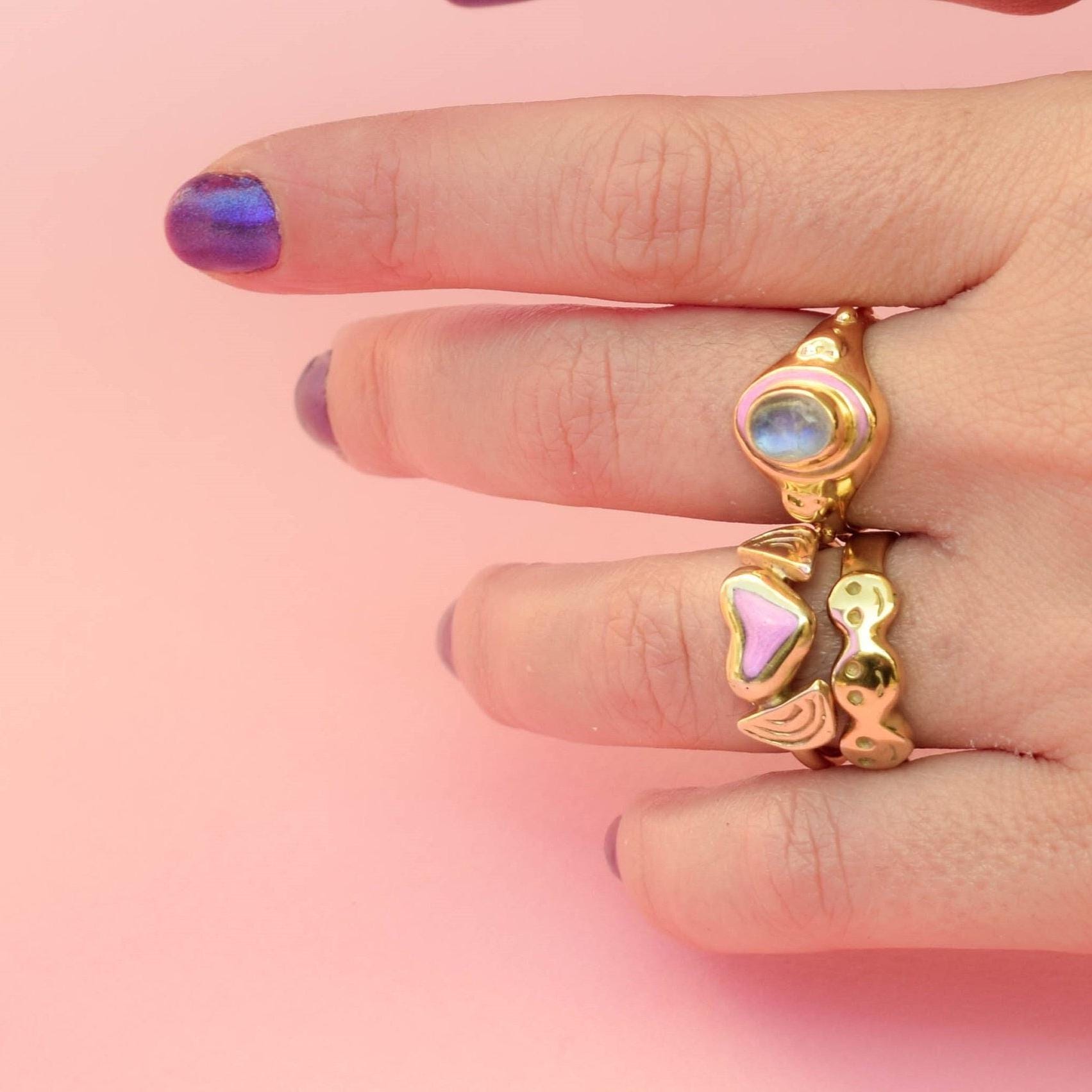 A hand wearing two rings, one with a bold heart design and another with a moonstone-like stone, on a pink background.