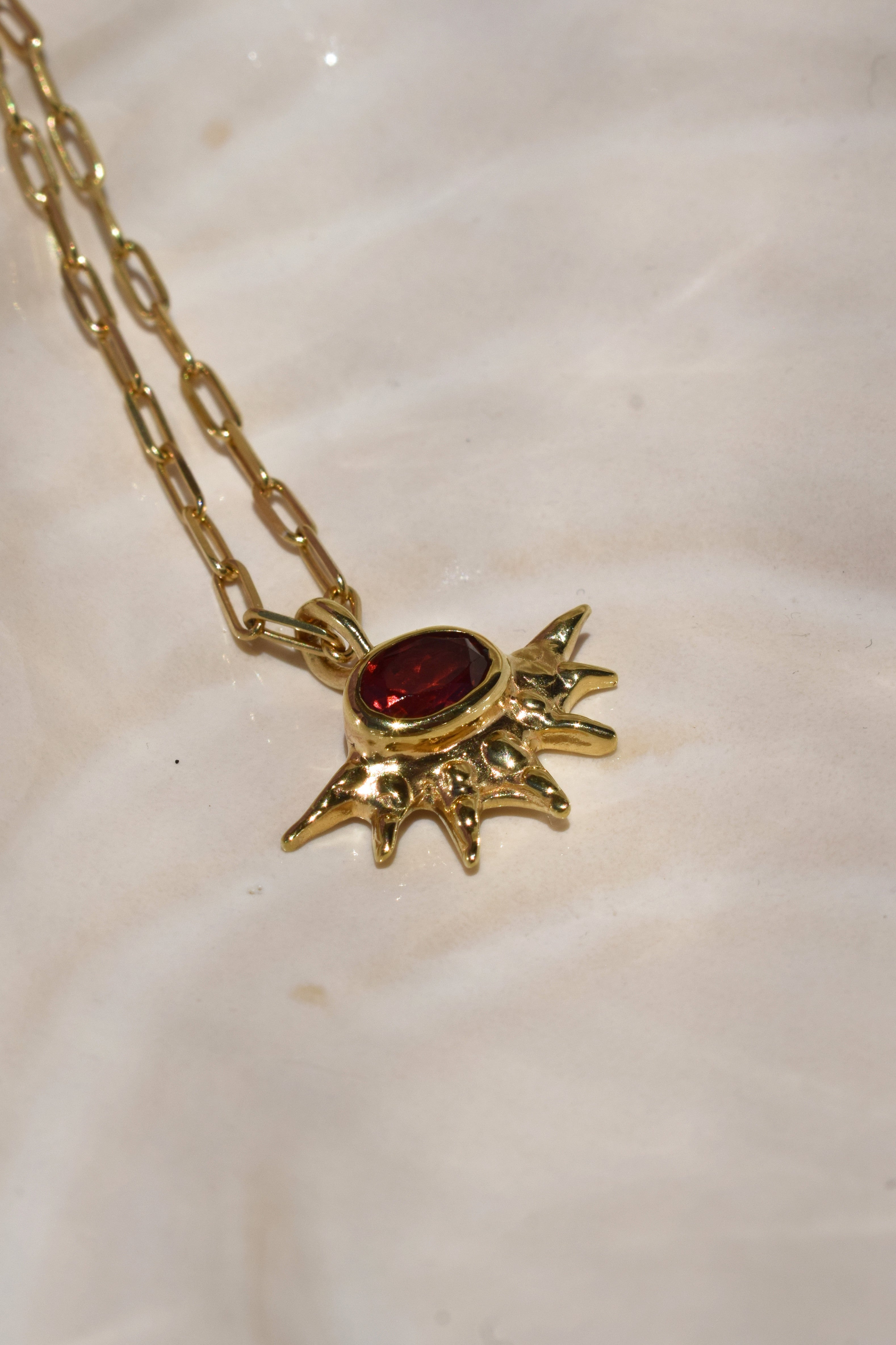 A radiant pendant on a paperclip chain, featuring a deep red garnet at the center of an asymmetrical, spiked gold setting. Bold and edgier than a traditional sunburst, it's easy to wear solo or layered.