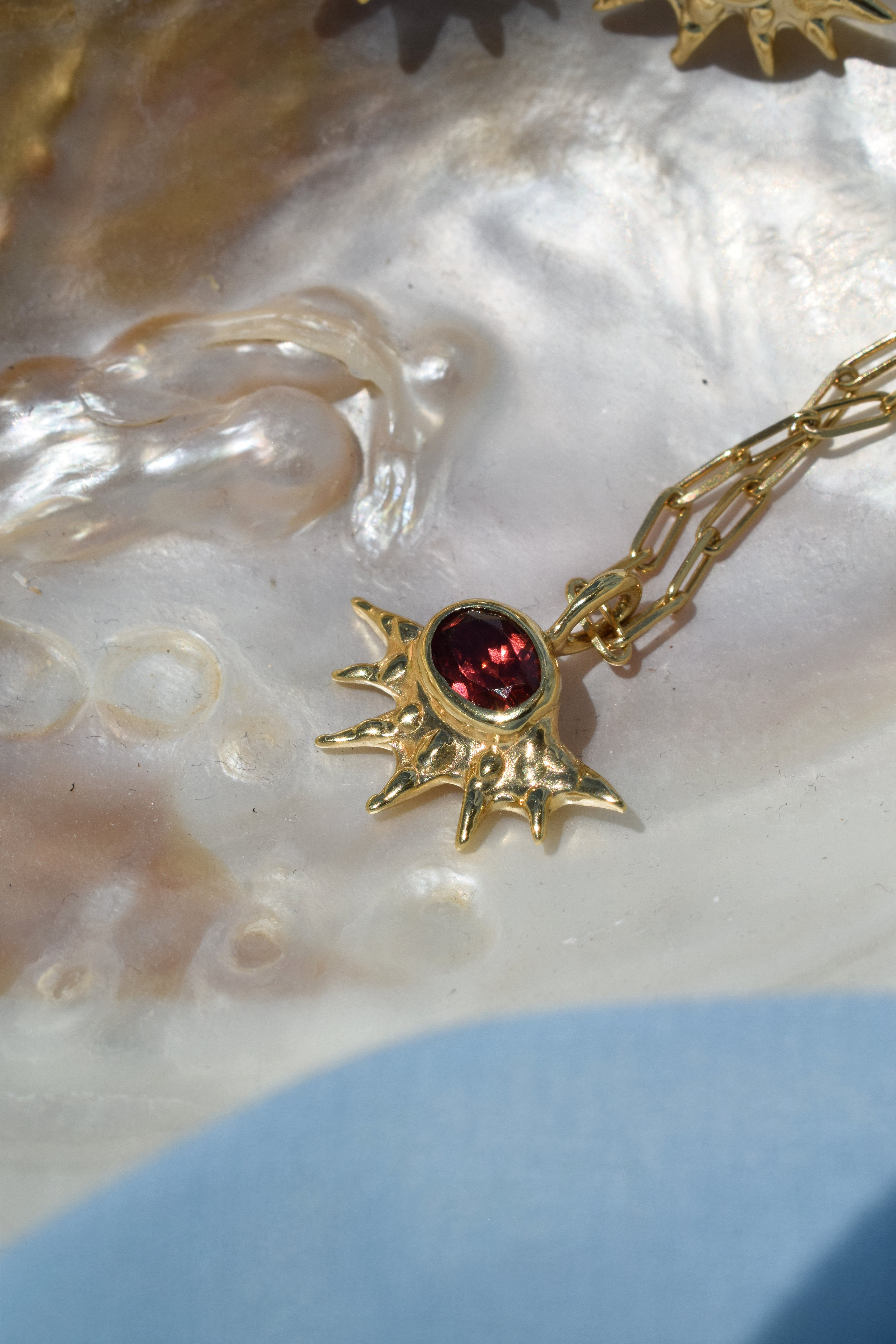 A radiant pendant on a paperclip chain, featuring a deep red garnet at the center of an asymmetrical, spiked gold setting. Bold and edgier than a traditional sunburst, it's easy to wear solo or layered.