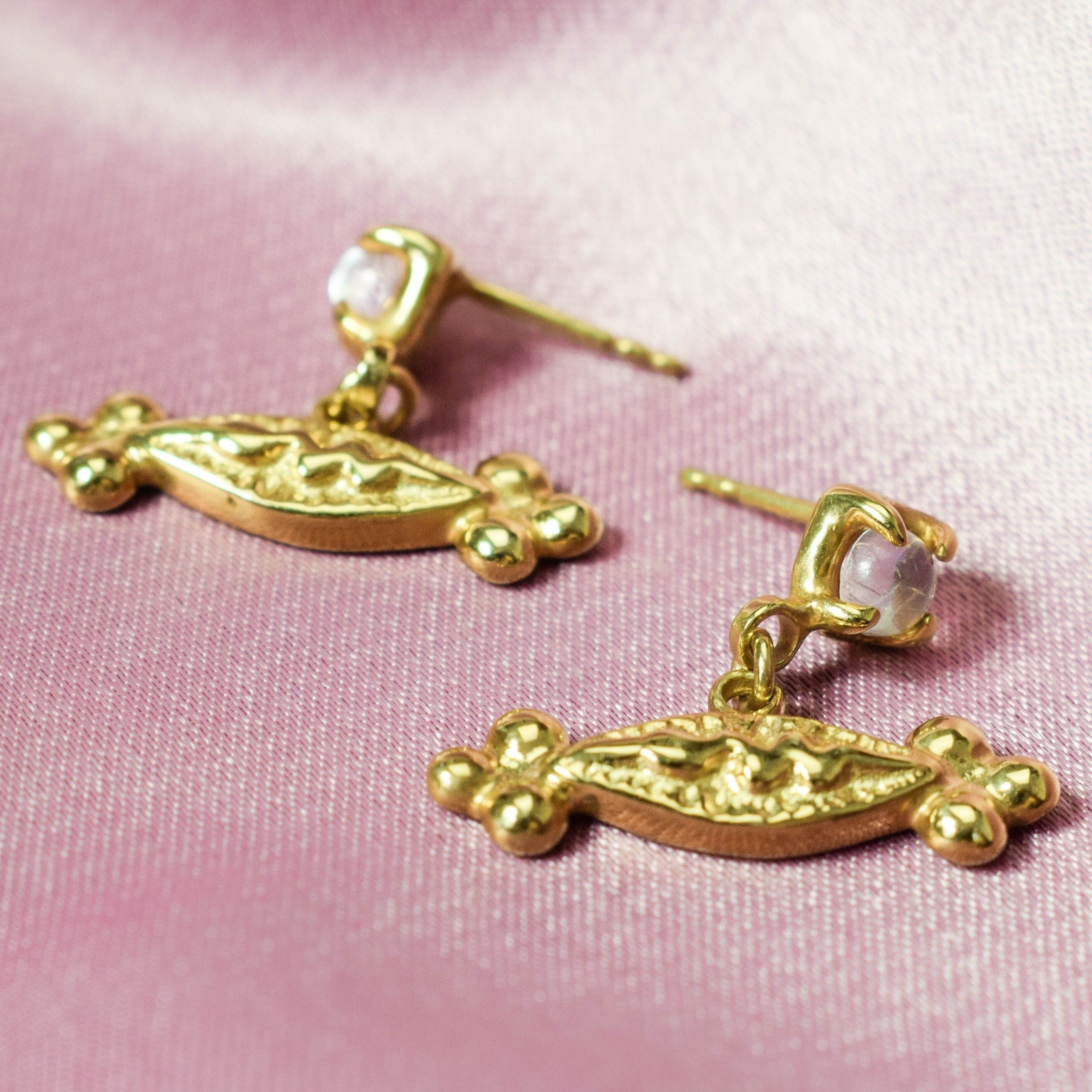 A pair of elegant stud earrings featuring gold-tone snake charms and moonstones, displayed against a pink background.