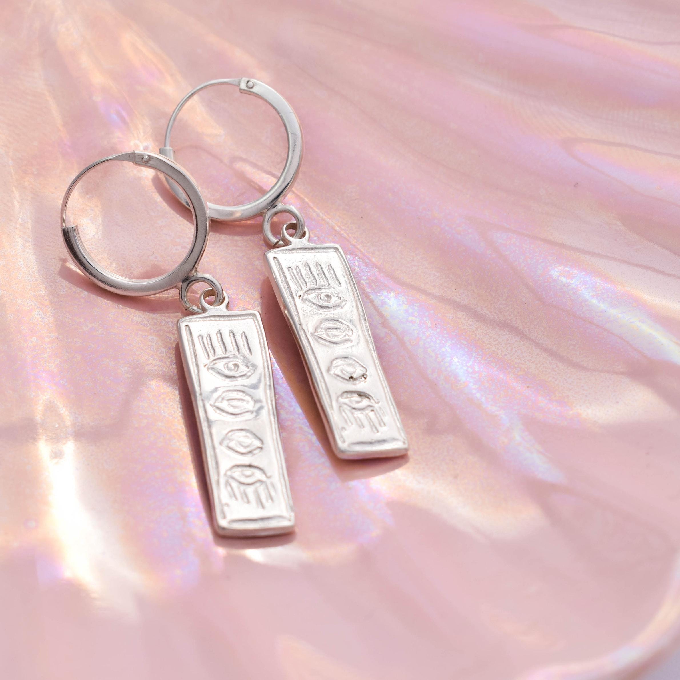 A pair of silver hoop earrings with vertical charm featuring an evil eye design, displayed on a pink iridescent surface.