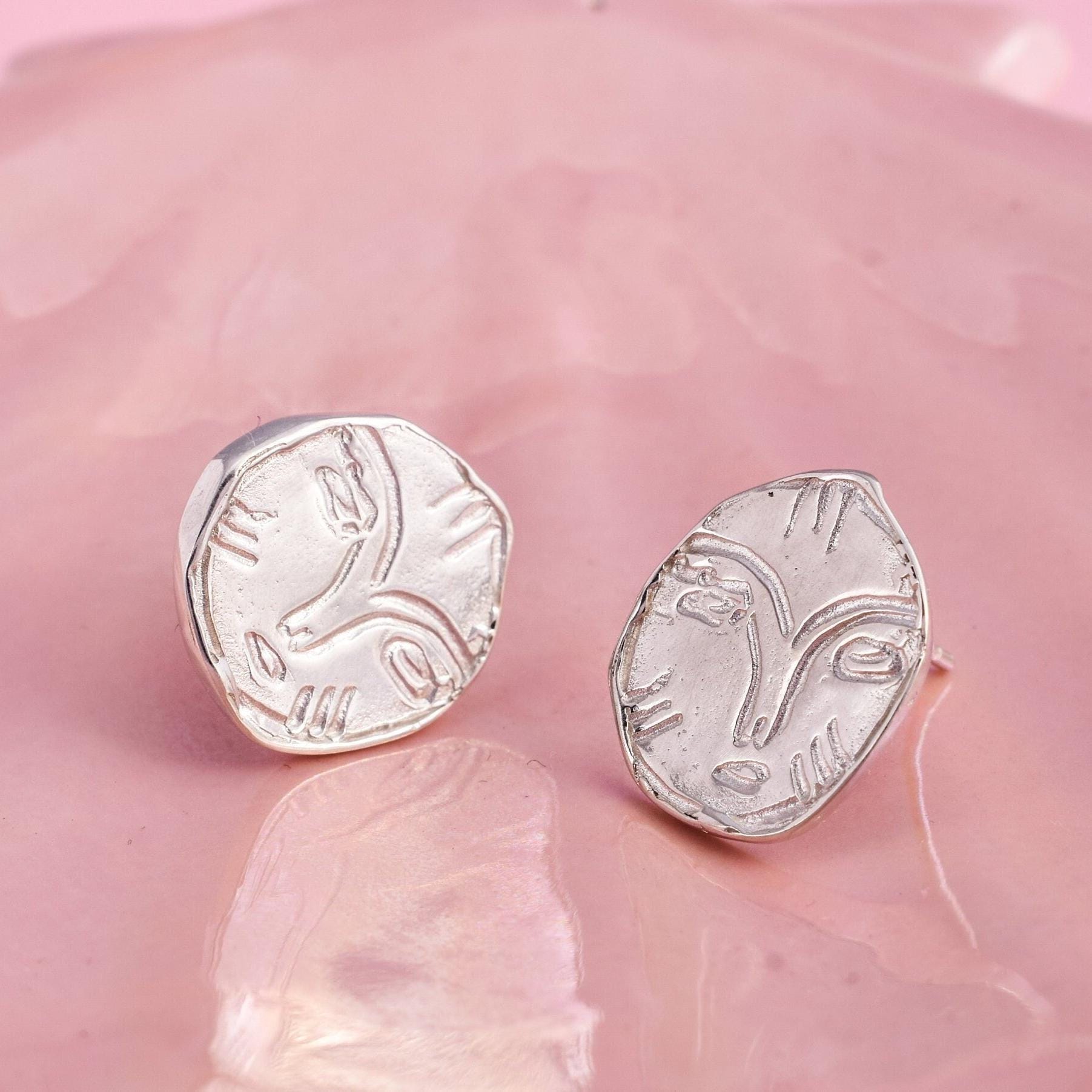 A pair of silver stud earrings, each featuring a unique face design, displayed against a pink background.
