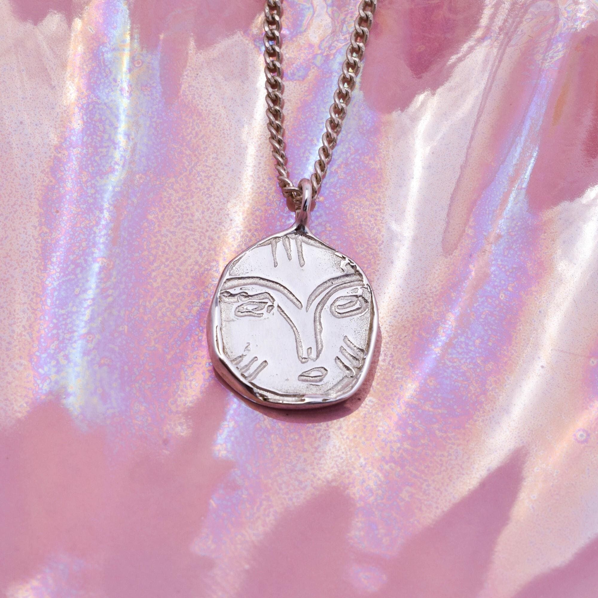 A silver necklace with a circular pendant featuring a face design, displayed against a pink holographic background.