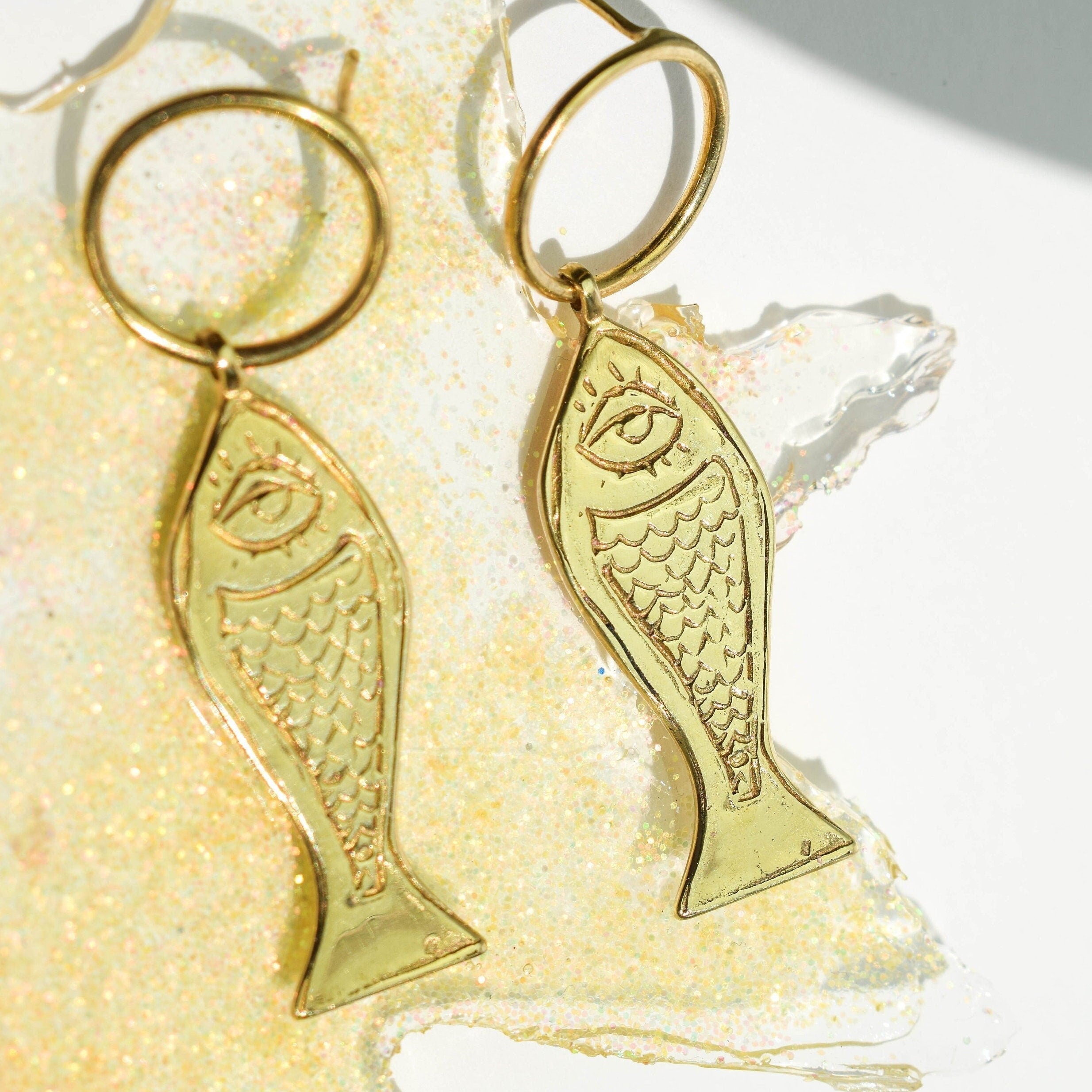 A pair of gold-tone fish earrings with human eyes, displayed against a reflective background.
