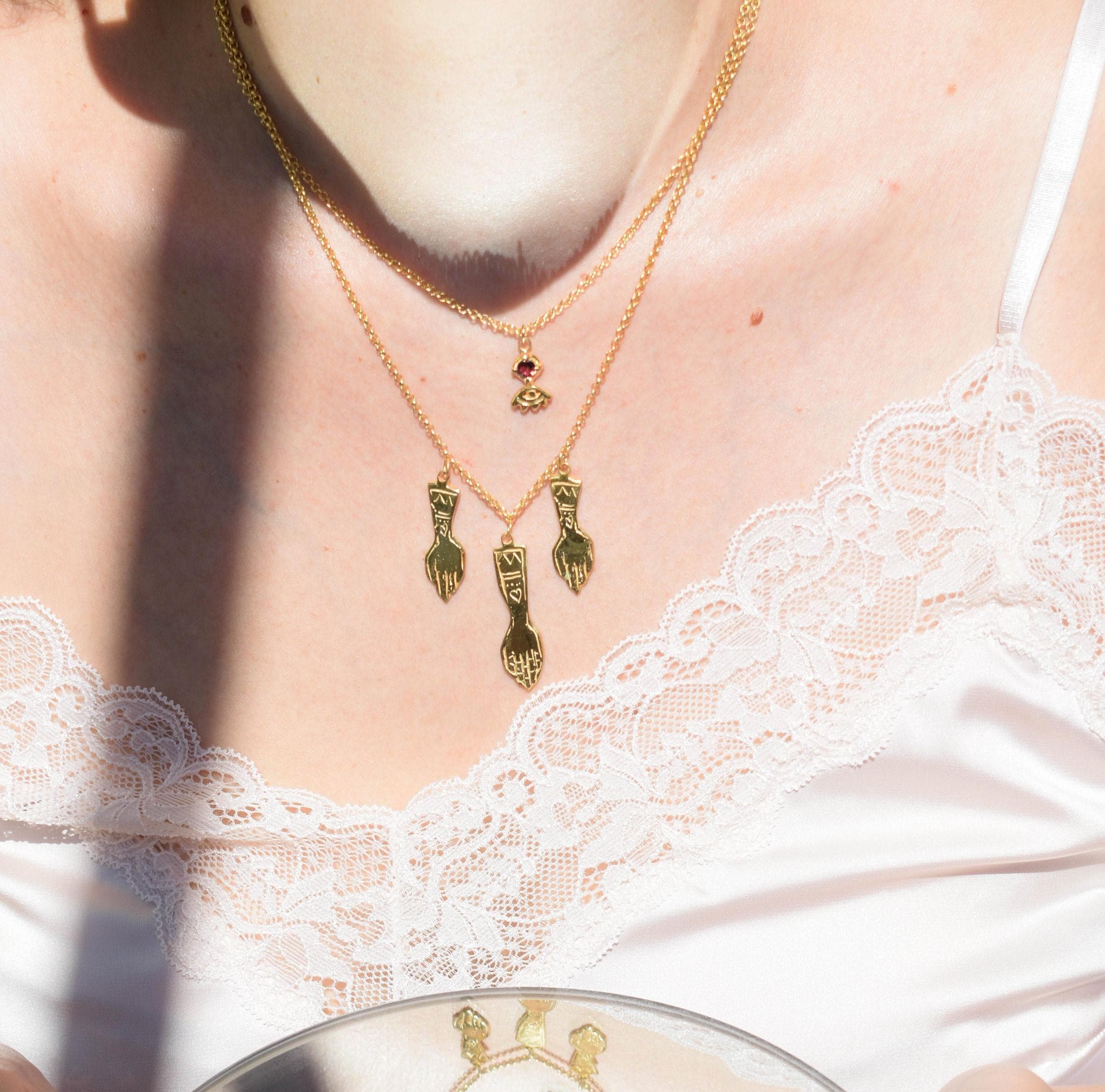A person wearing a gold-tone witch necklace with hand charms, layered with other necklaces.