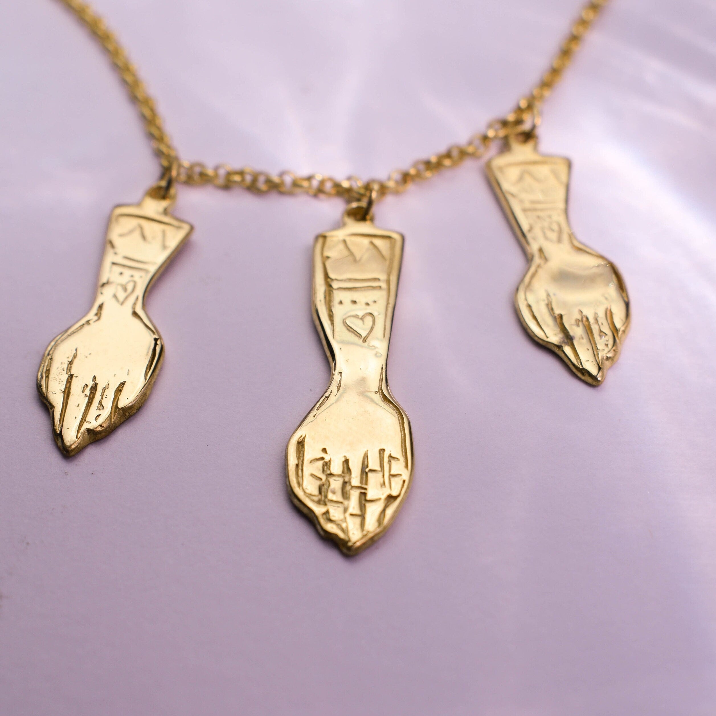 A gold-tone necklace featuring three pendant charms shaped like hands with hearts on them, displayed against a light purple background.