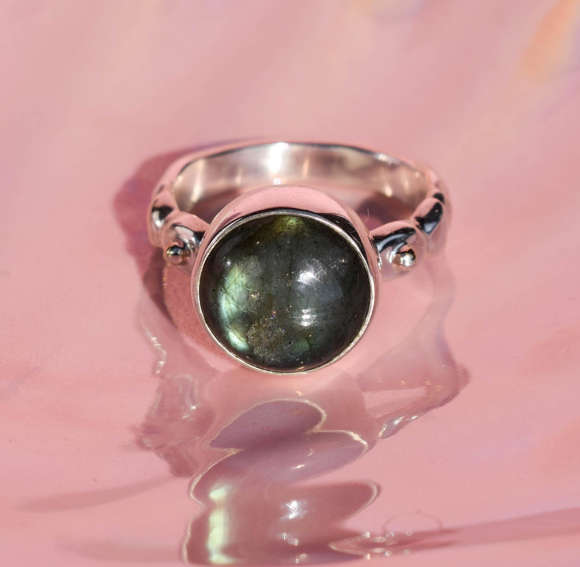 A labradorite ring with a silver band on a reflective surface, the gemstone displays shades of green with a flash of light.