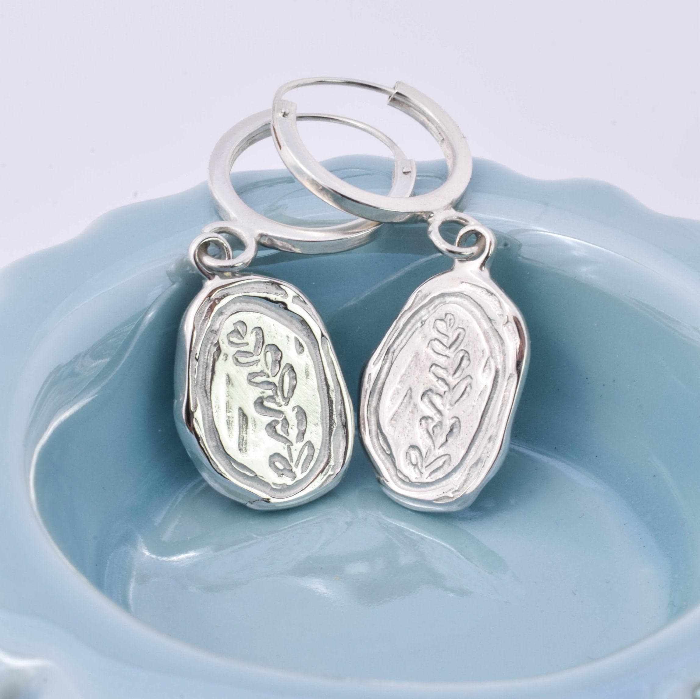 A pair of silver hoop earrings with leaf charms attached, displayed in a blue dish.