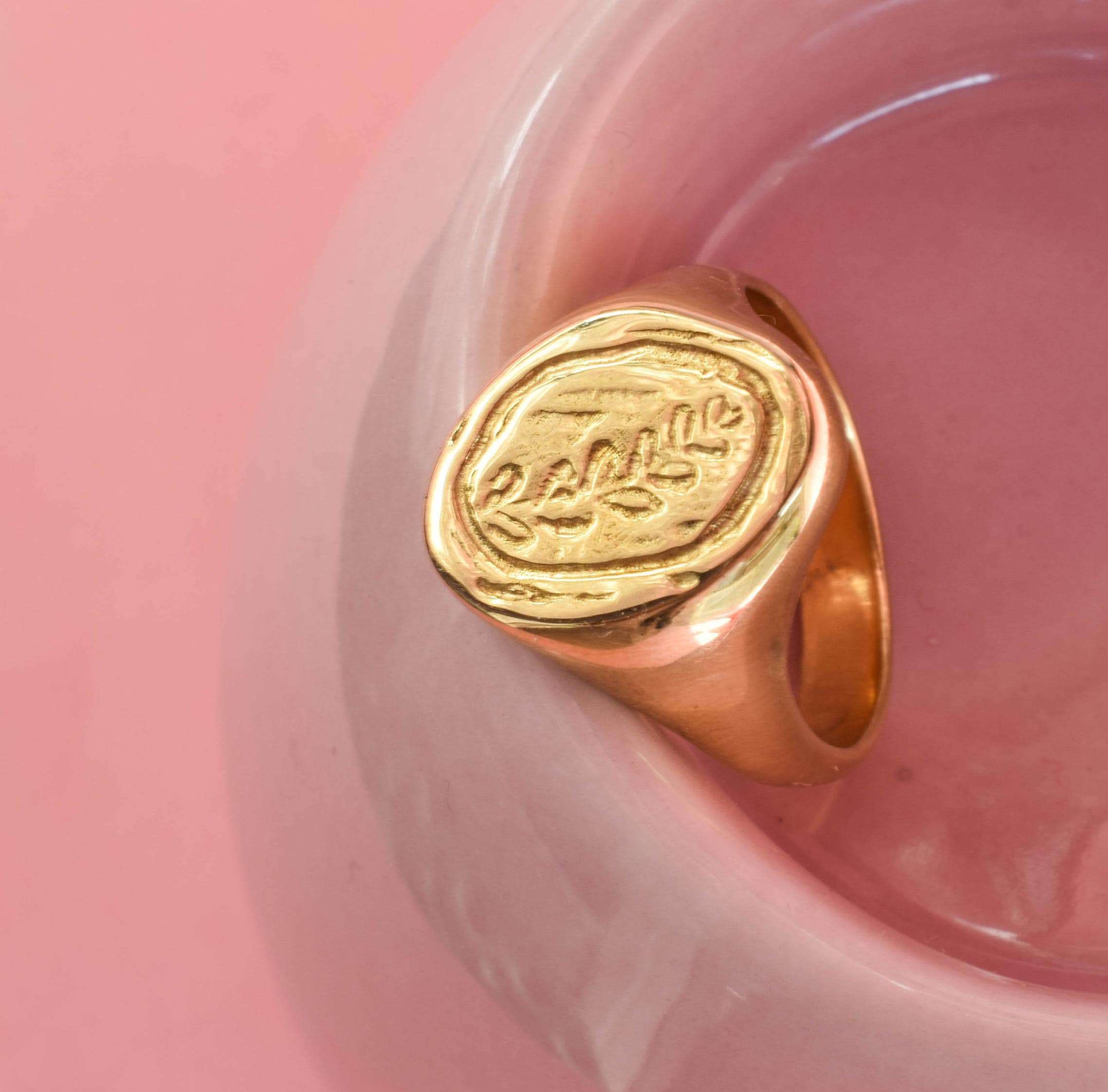 A gold-colored signet ring with a leaf design, placed on a pink surface.