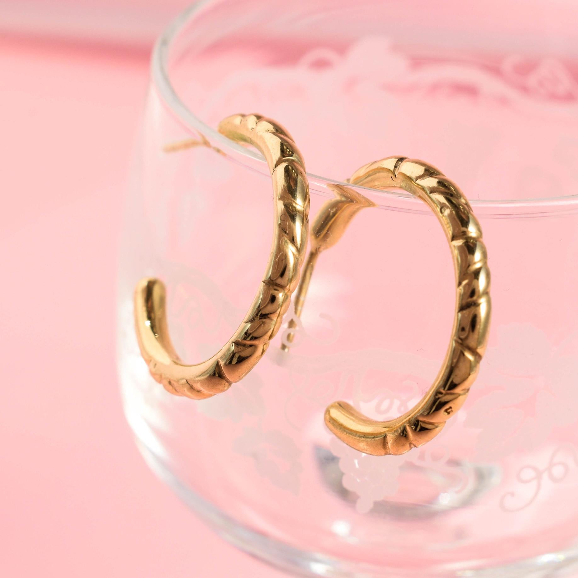 A pair of medium-sized gold-colored stacking hoop earrings displayed on a clear glass surface with a pink background.