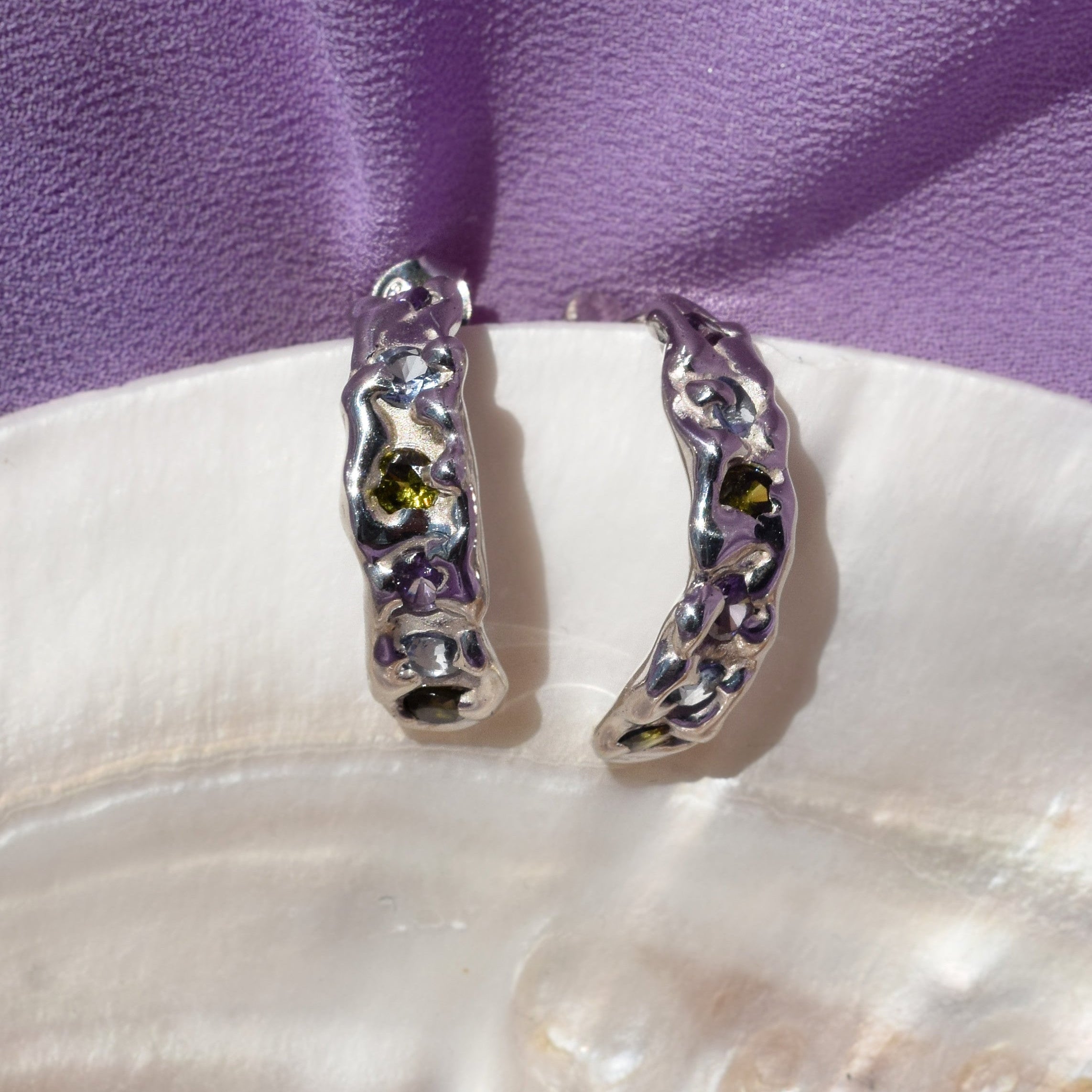A pair of silver hoop earrings with textured organic edges and small purple, blue and green gemstones, displayed on a white shell against a purple fabric background.