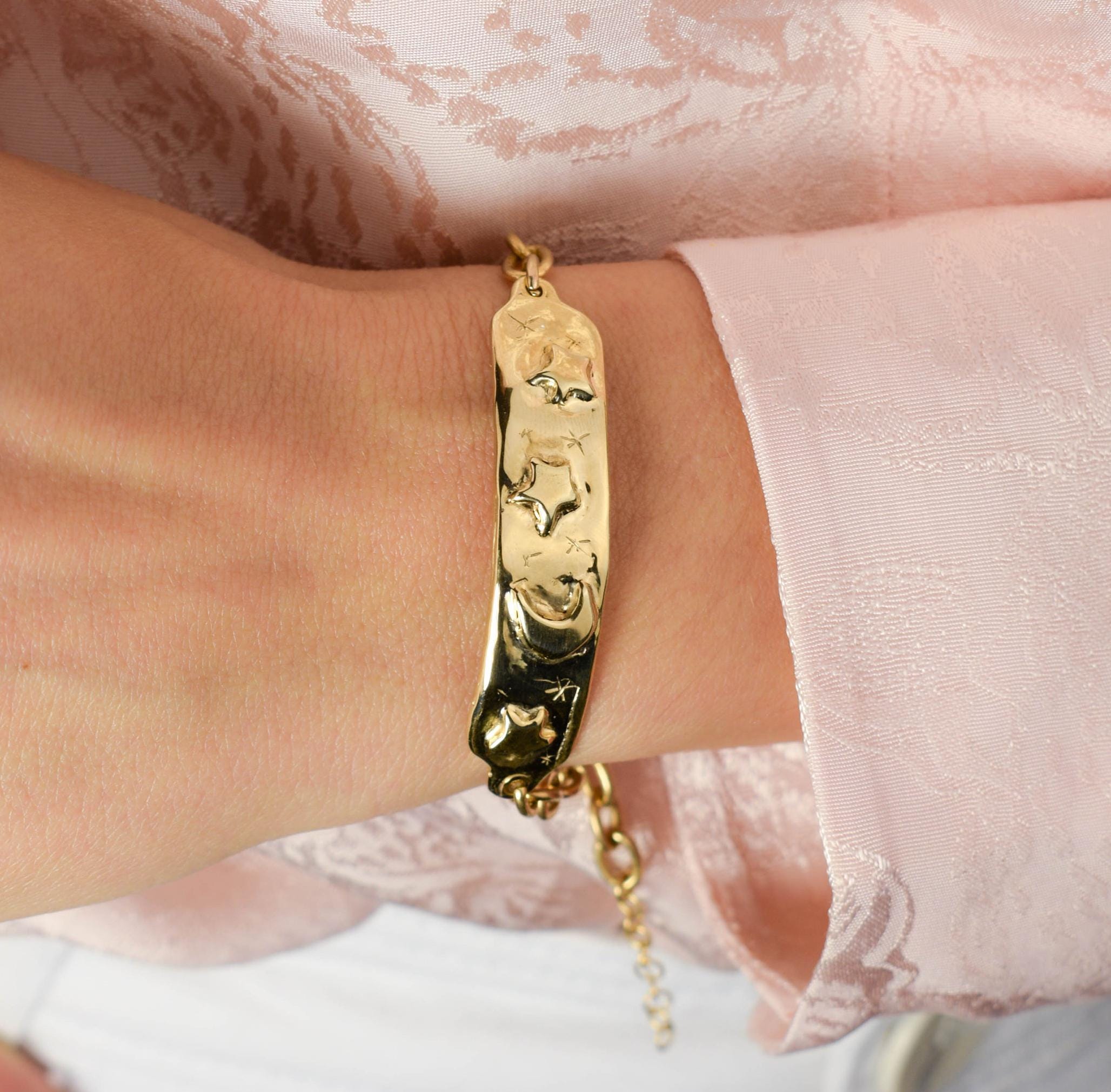 A person's wrist wearing a gold plated tag bracelet with three stars and a shiny moon, against a pink background.