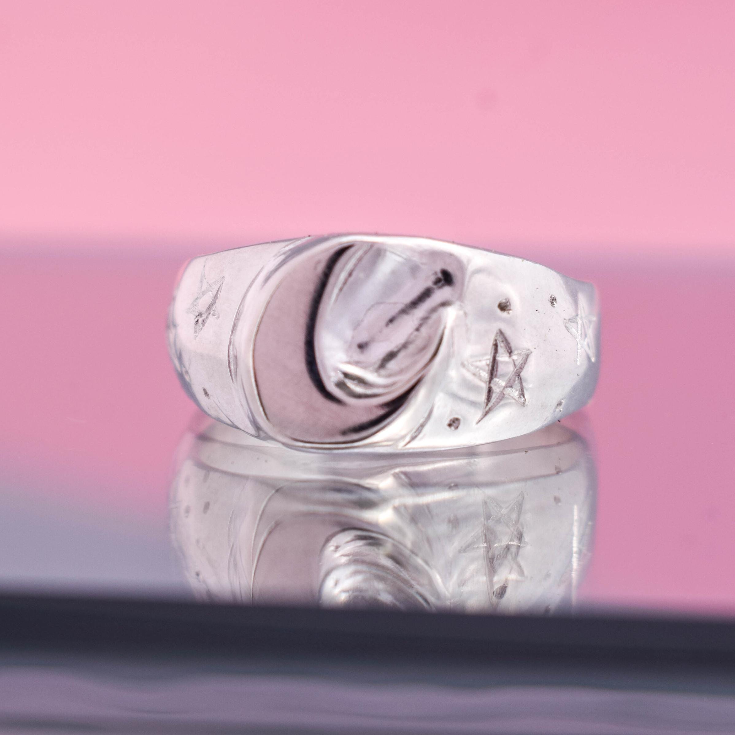 A sterling silver signet ring with a crescent moon design set against a pink background.