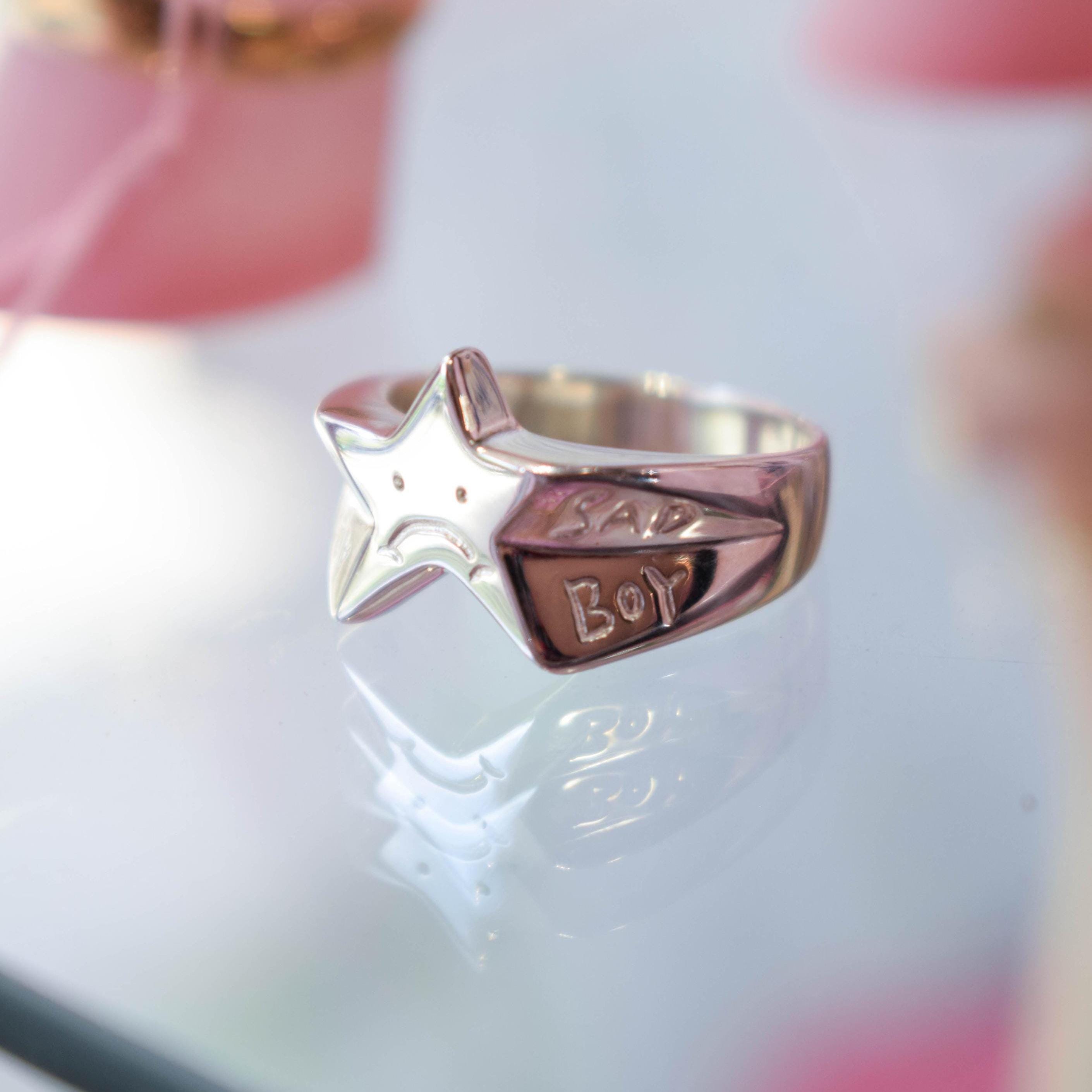 A sterling silver signet ring featuring a star shape with a sad face design on it, displayed on a reflective surface.