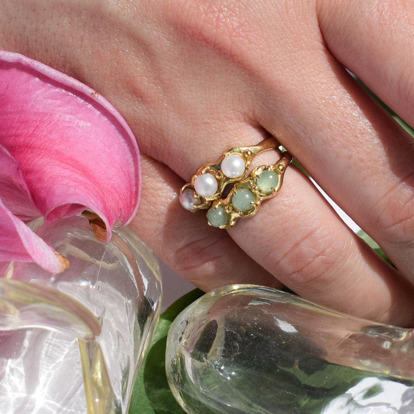 A gold-colored ring with alternating white and green stones on a person's hand, with a pink flower and glass in the background.