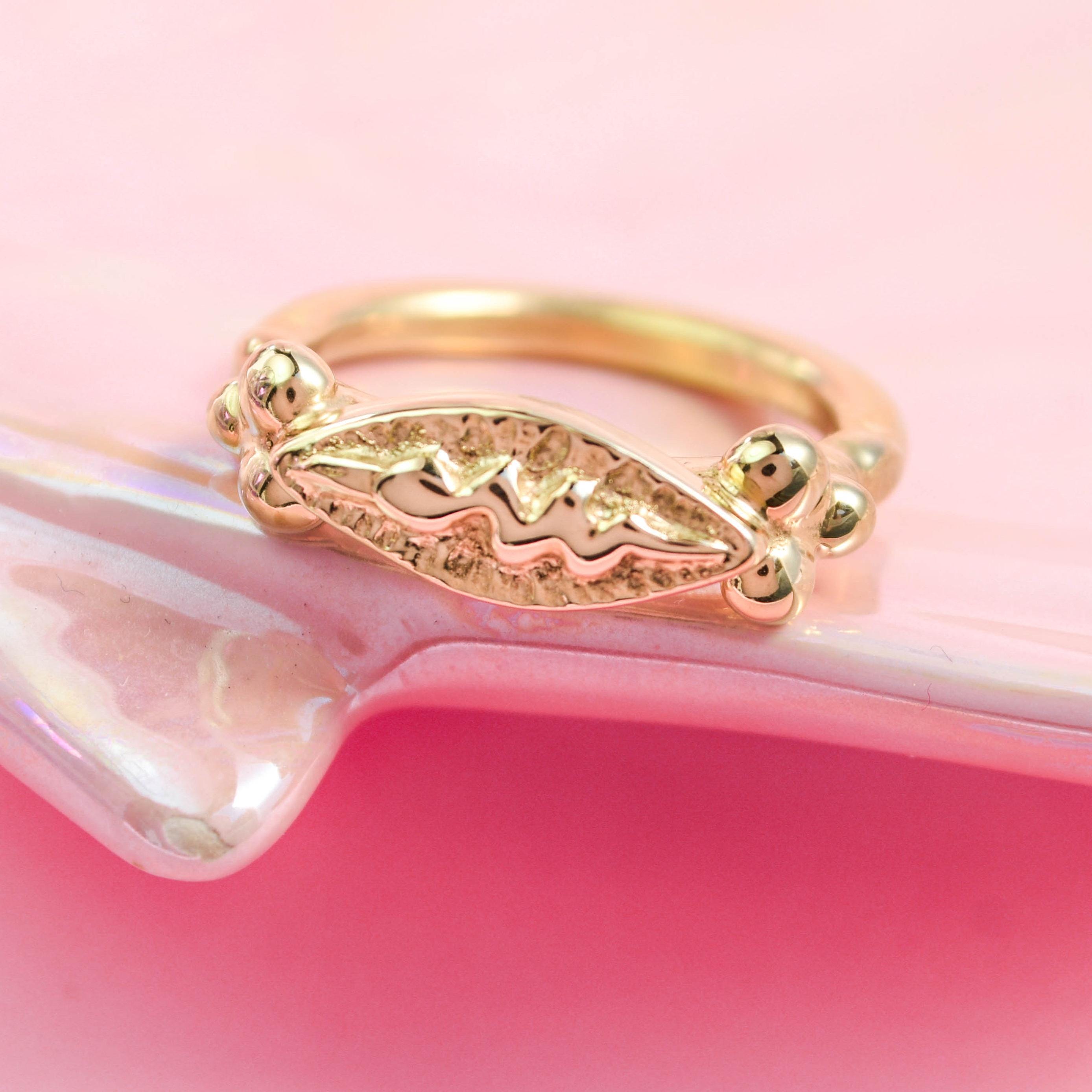 A gold-colored snake ring on a pink surface, featuring intricate detailing and a shiny finish.