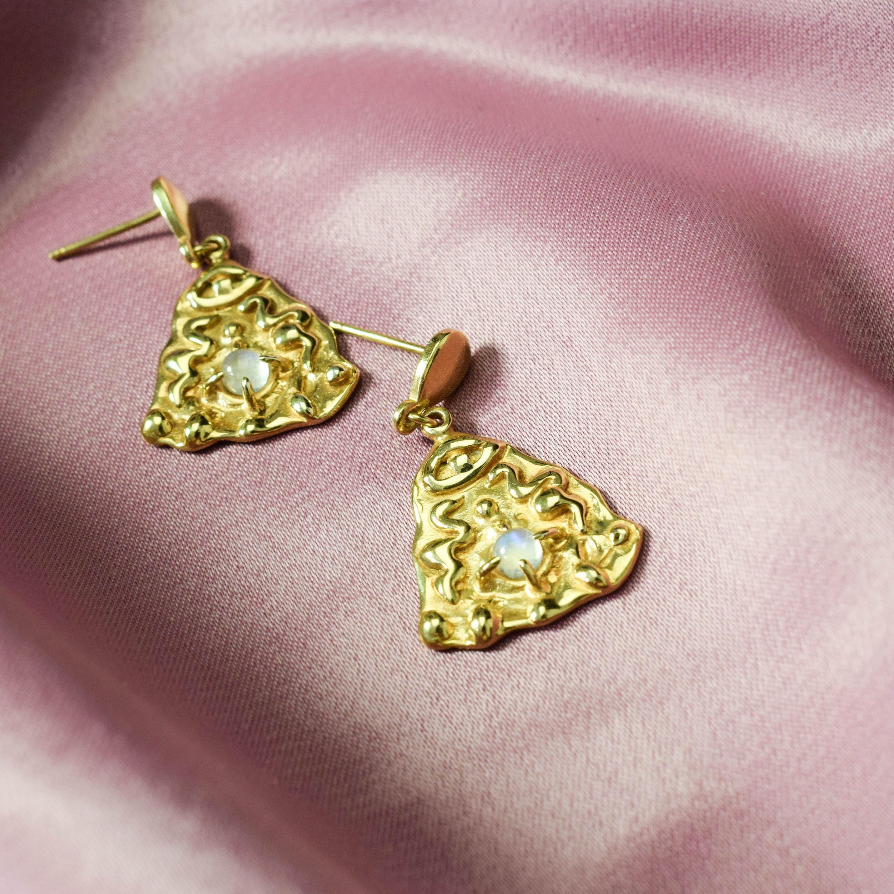 A pair of gold-tone earrings featuring a snake motif with eyes and a moonstone gemstone, displayed on a pink satin background.